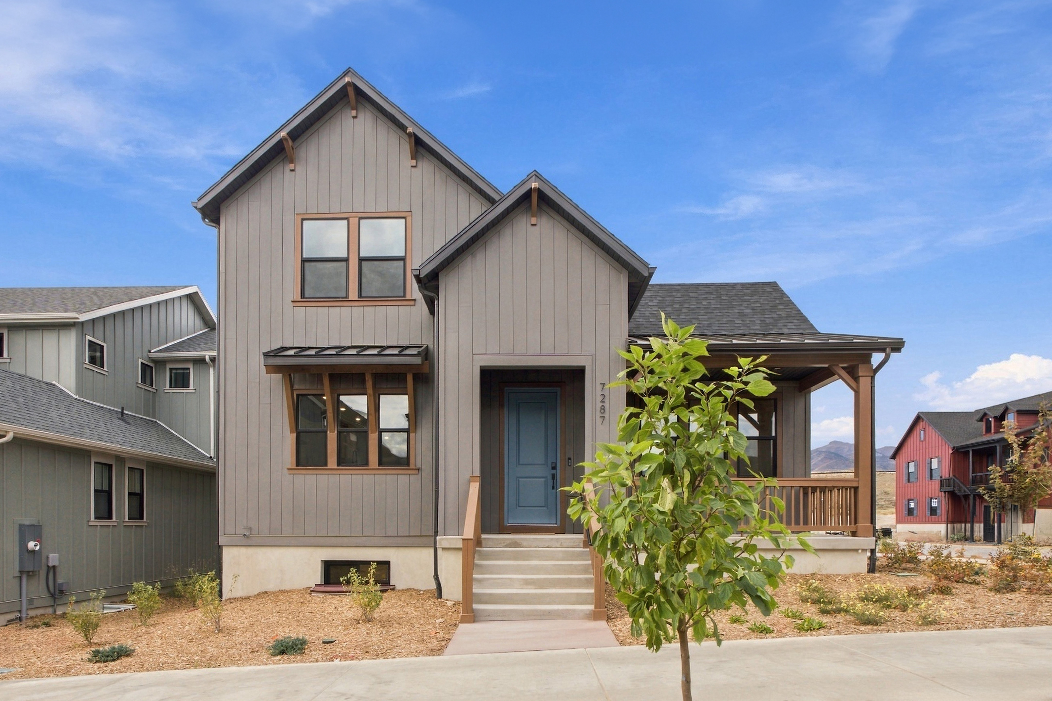 A small green tree and a concrete sidewalk sit in the foreground of a modern grey house under a bright blue sky.