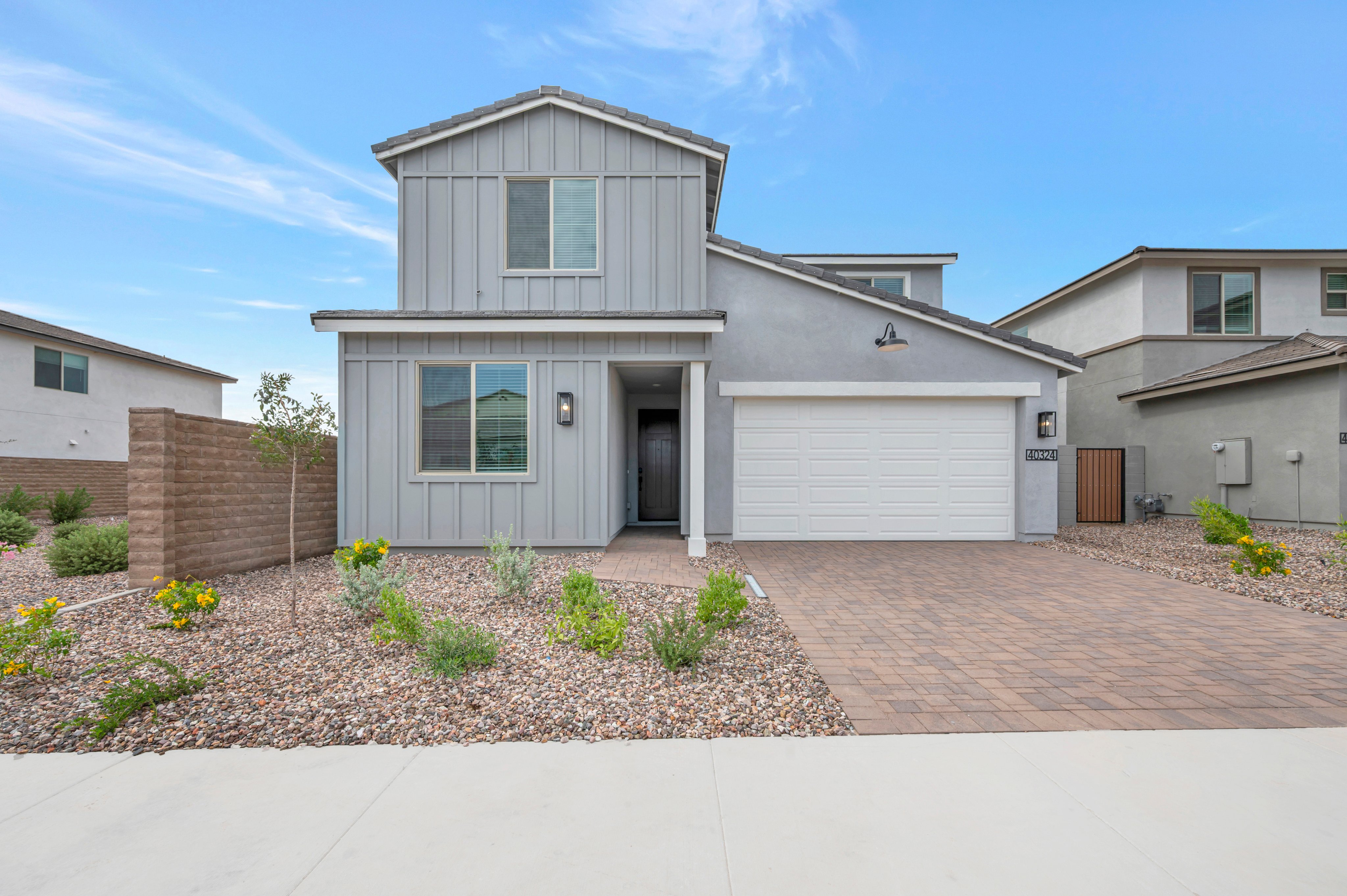A modern grey two-story house with a white garage door sits behind a paved driveway and gravel landscaping under a clear blue sky.