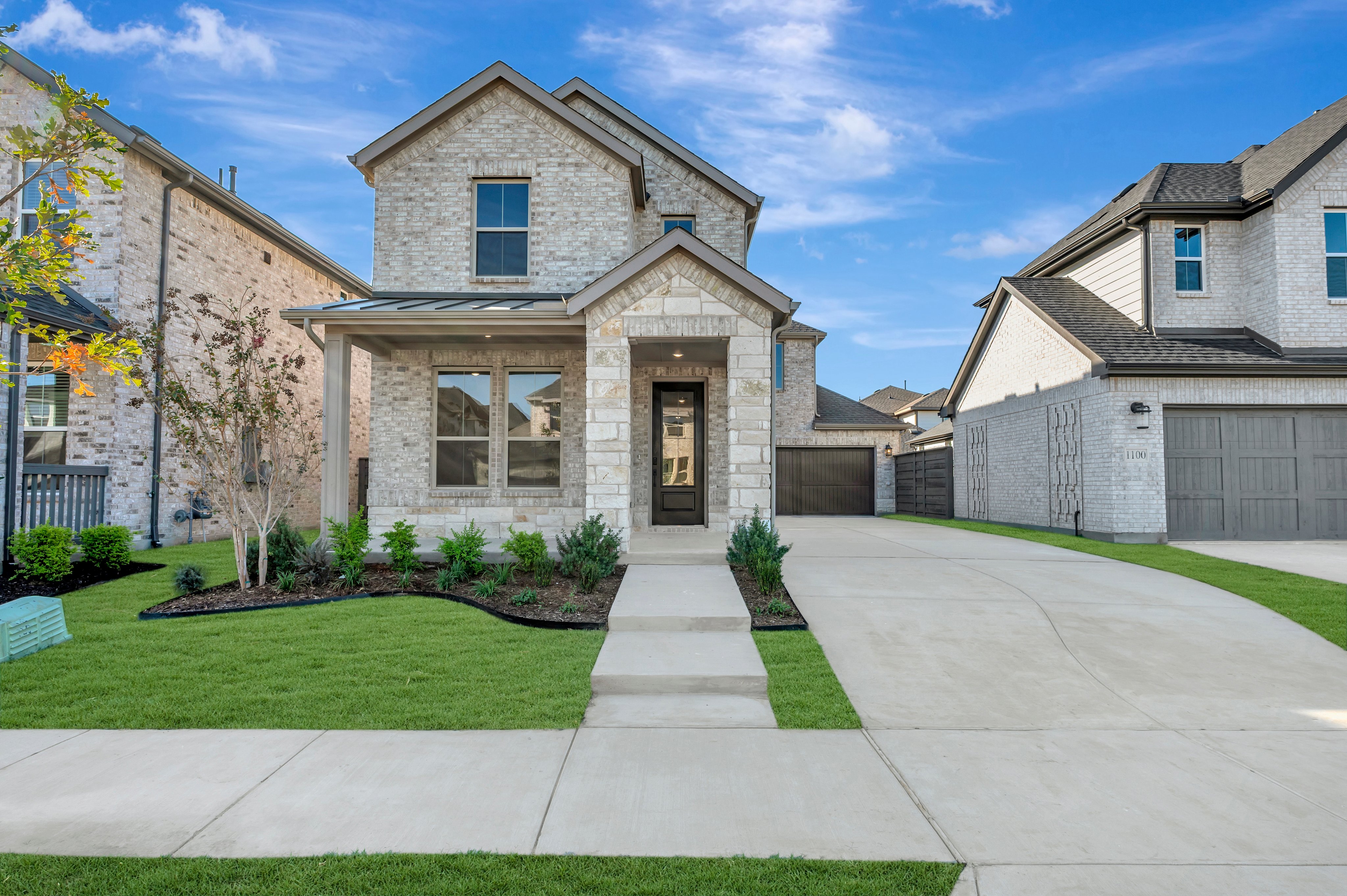 A modern light-colored stone house with a manicured green lawn and concrete driveway sits under a bright blue sky between two neighboring homes.