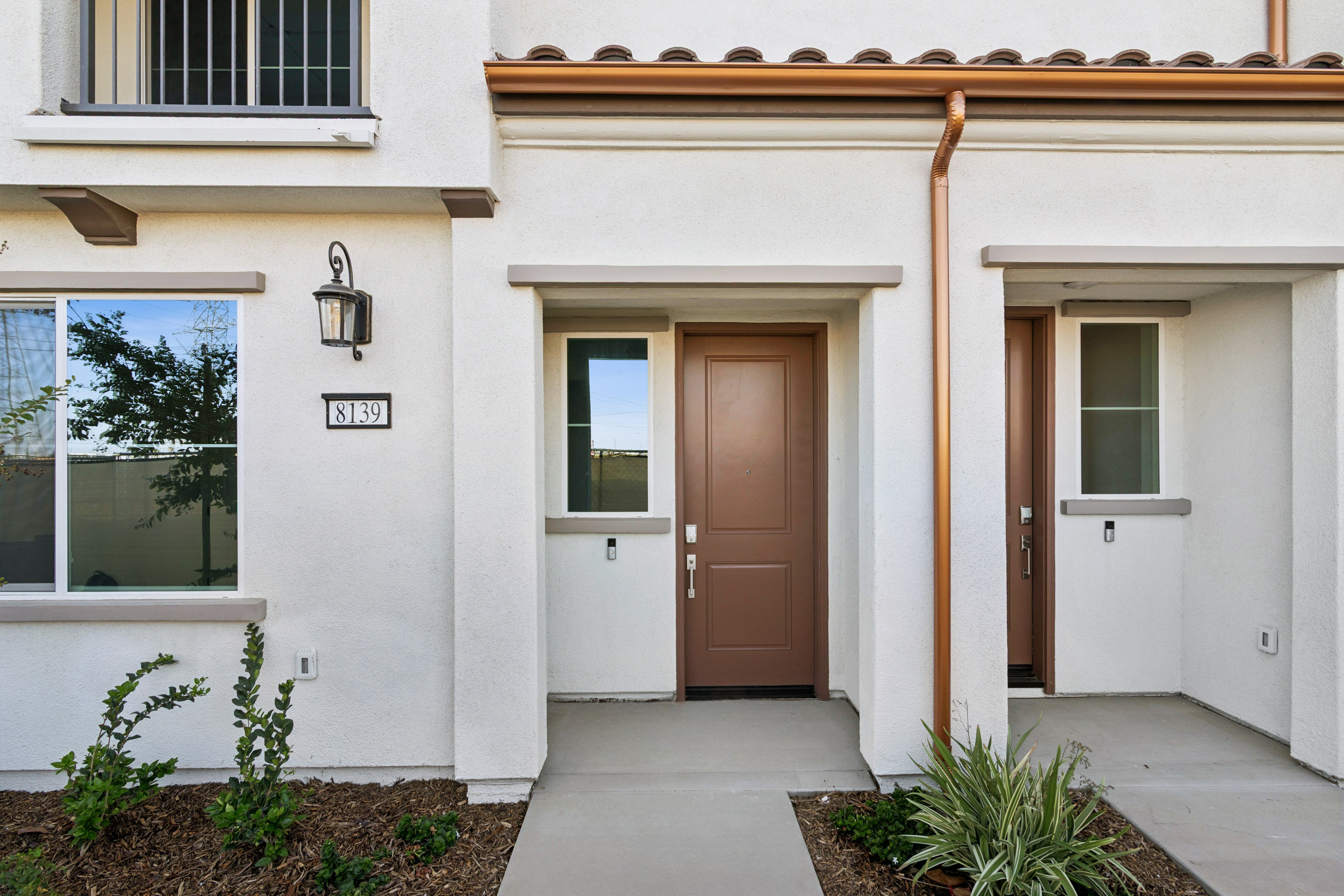 A brown front door and concrete walkway are in the foreground of a white stucco building with a copper gutter in the background.