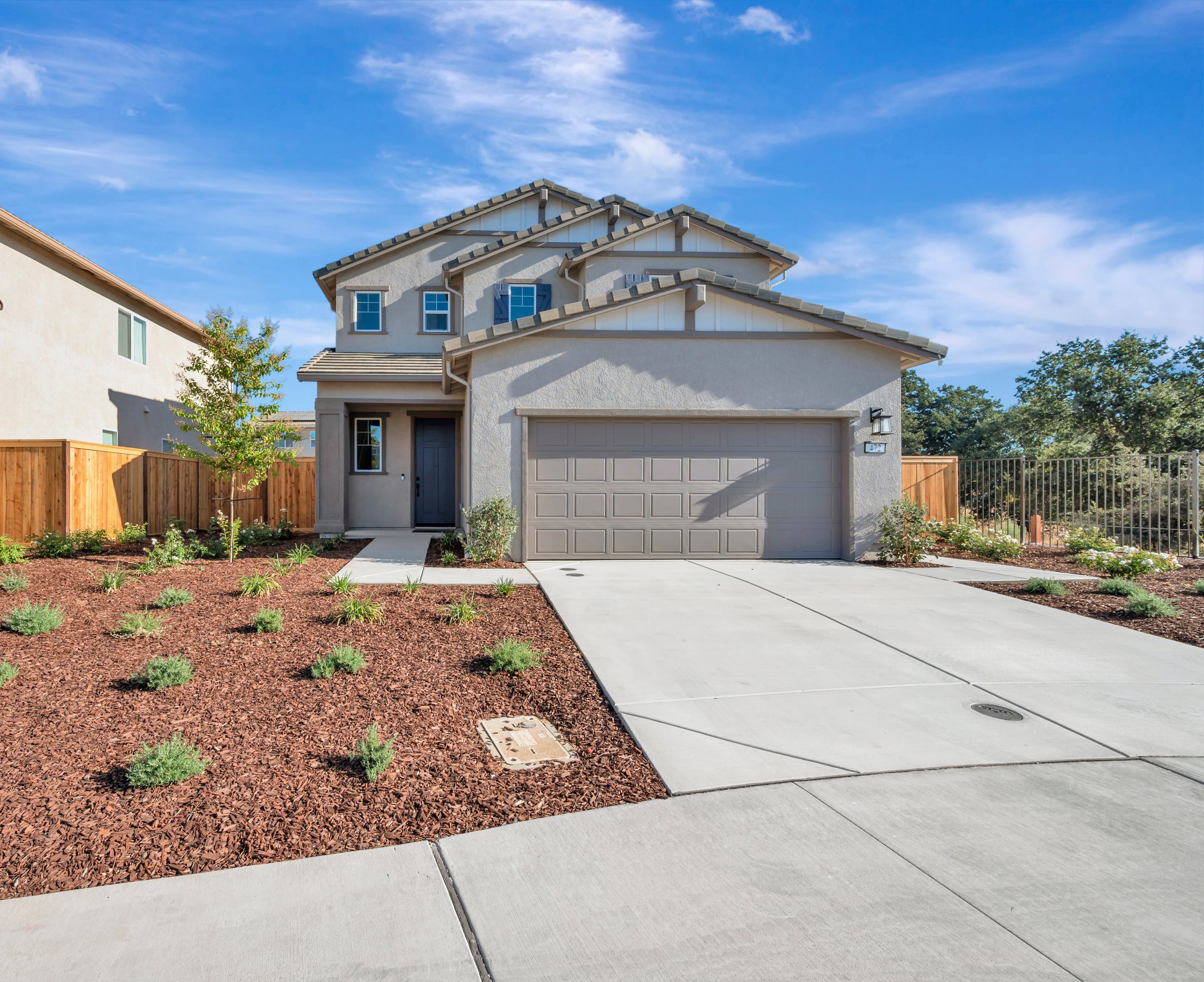 A modern two-story house with a grey garage door sits behind a concrete driveway and landscaped mulch beds under a bright blue sky.
