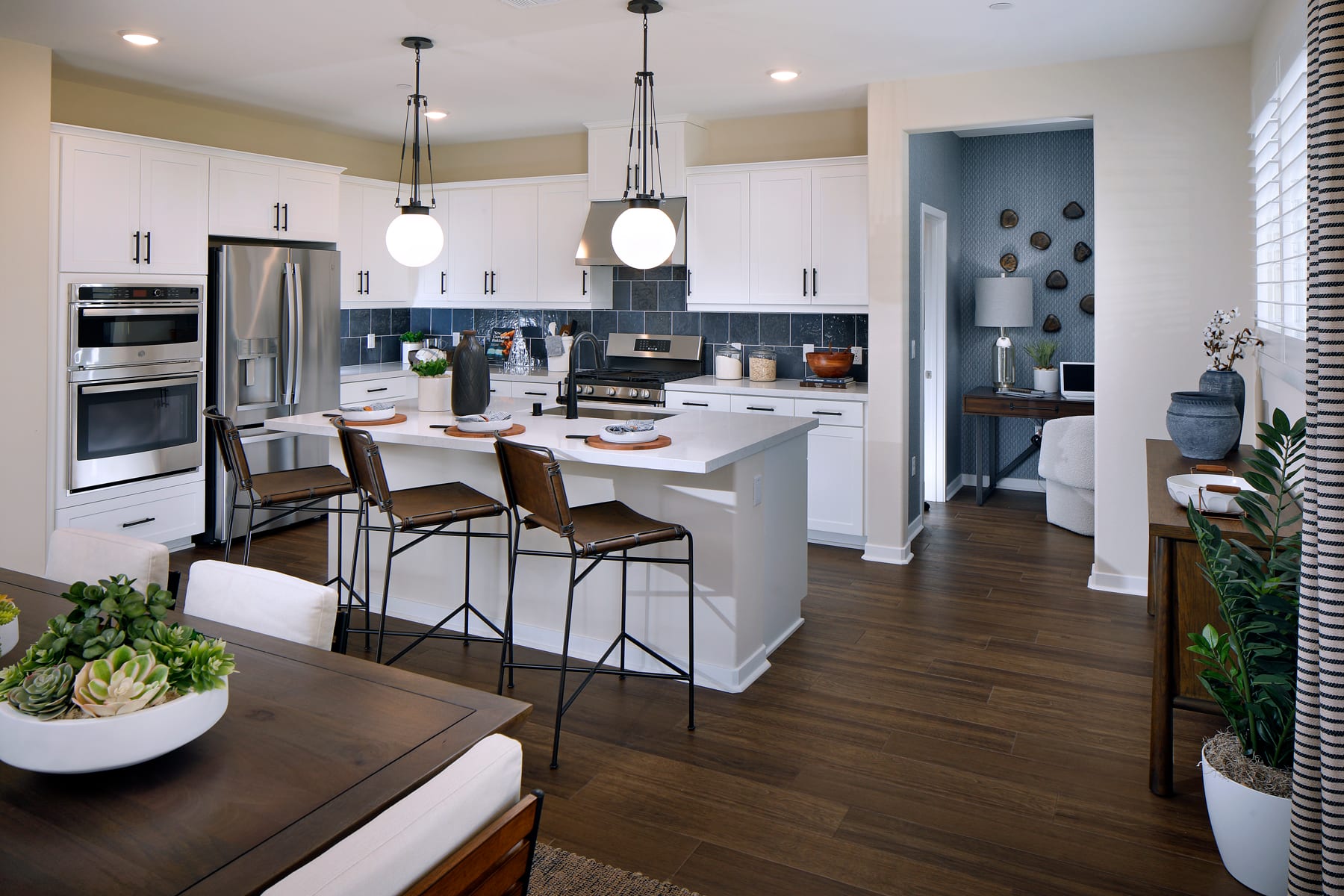 A modern, open-concept kitchen with white cabinets, stainless steel appliances, and a central island with bar stools, surrounded by a cozy living area with a brown sofa and potted plants.