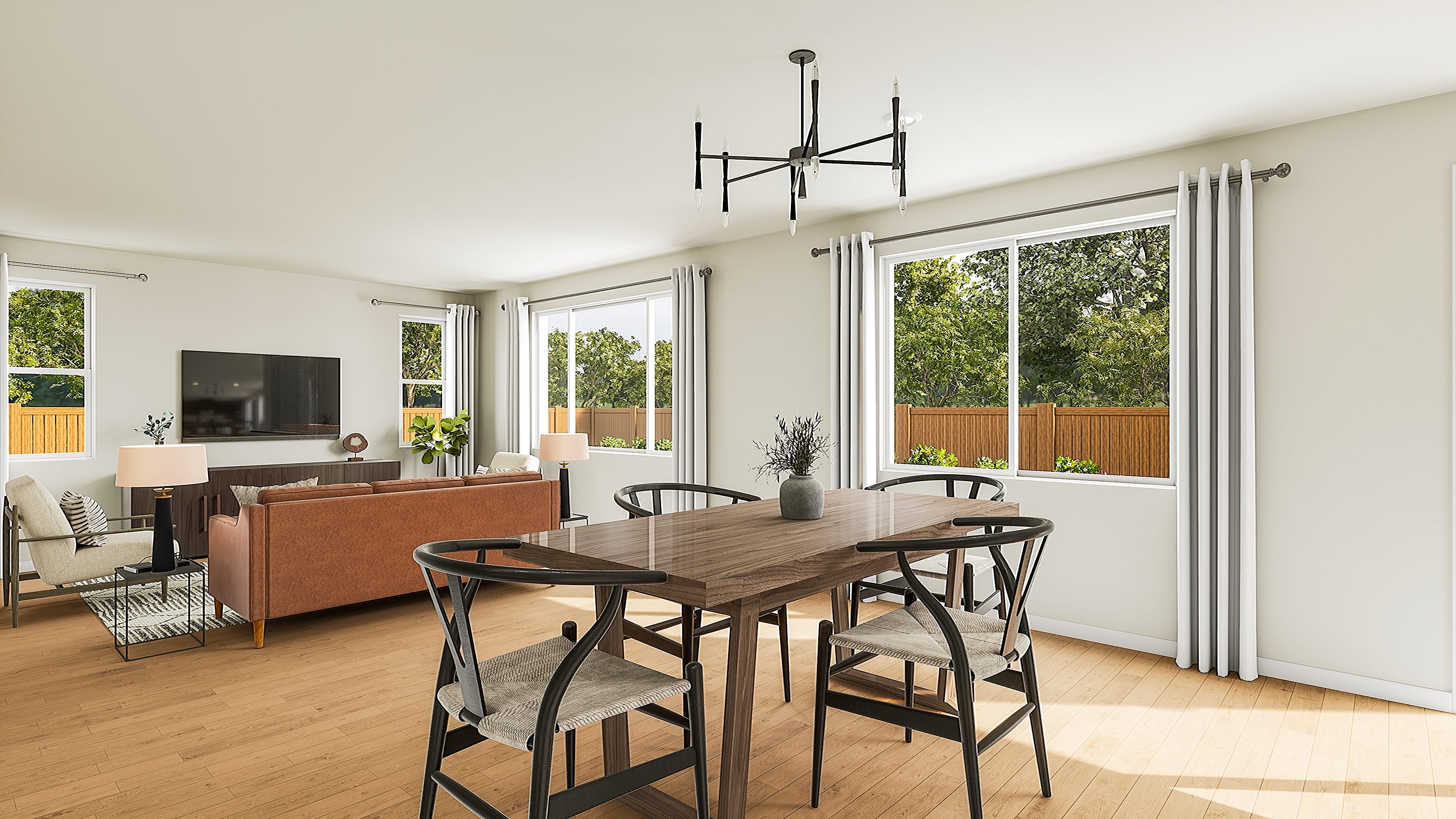 A modern dining room with a wooden table and chairs in the foreground is adjacent to a living room with a sofa and television, all bathed in natural light from large windows overlooking a green backyard.
