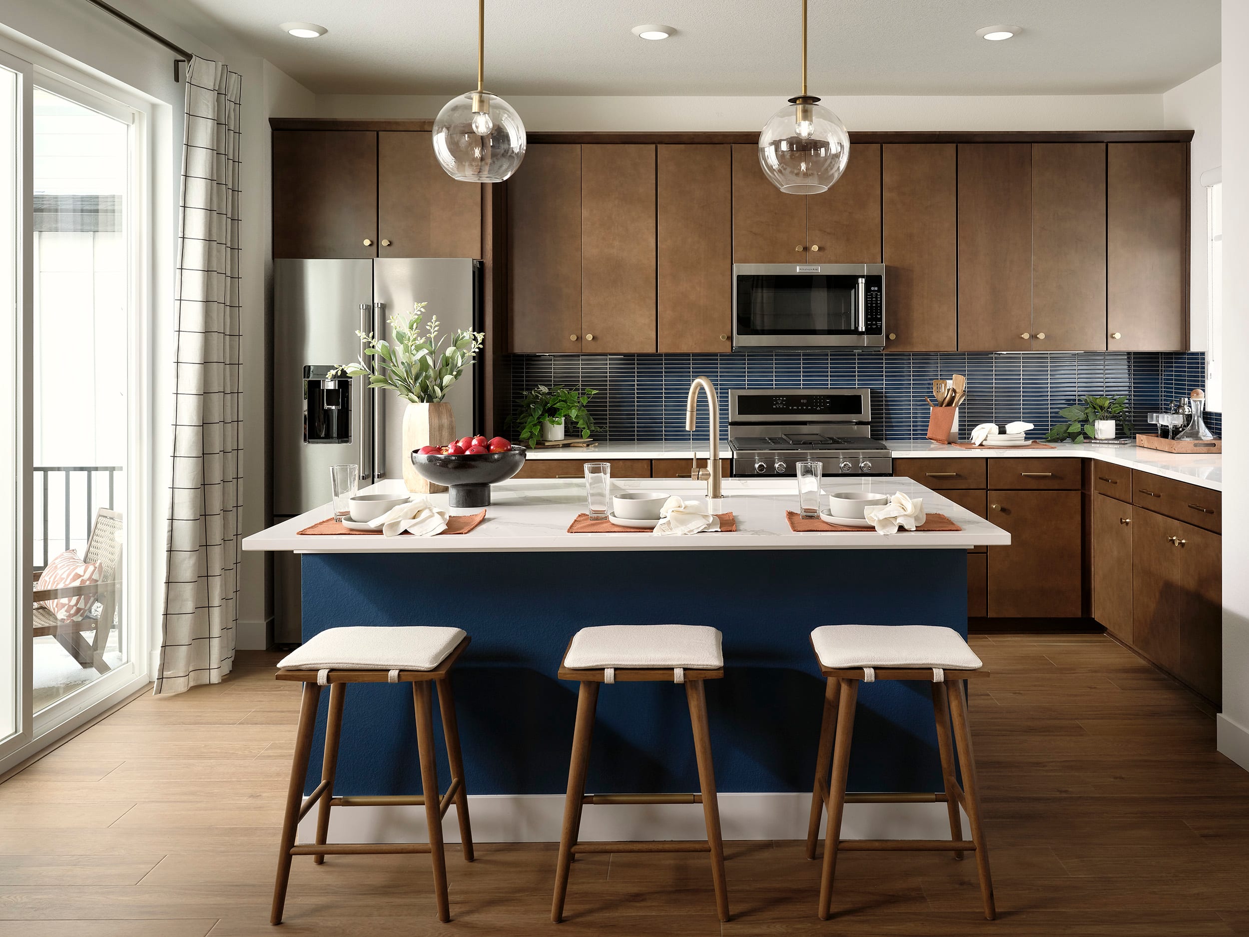 A modern and well-designed kitchen with a large island, wooden cabinets, and pendant lighting fixtures, set against a backdrop of large windows and a balcony.