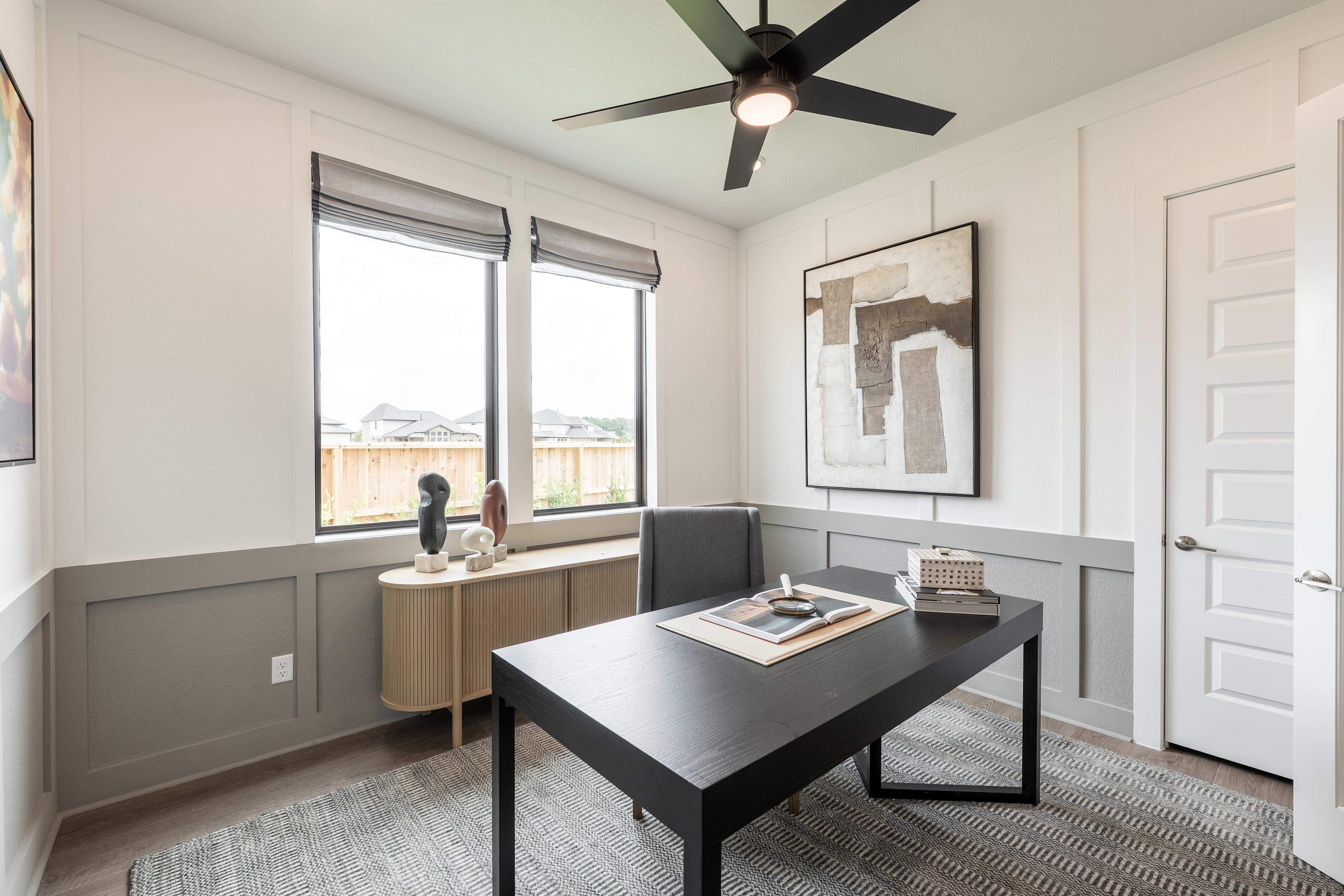 A modern and minimalist home office space with a black desk, a ceiling fan, and a framed artwork on the wall.