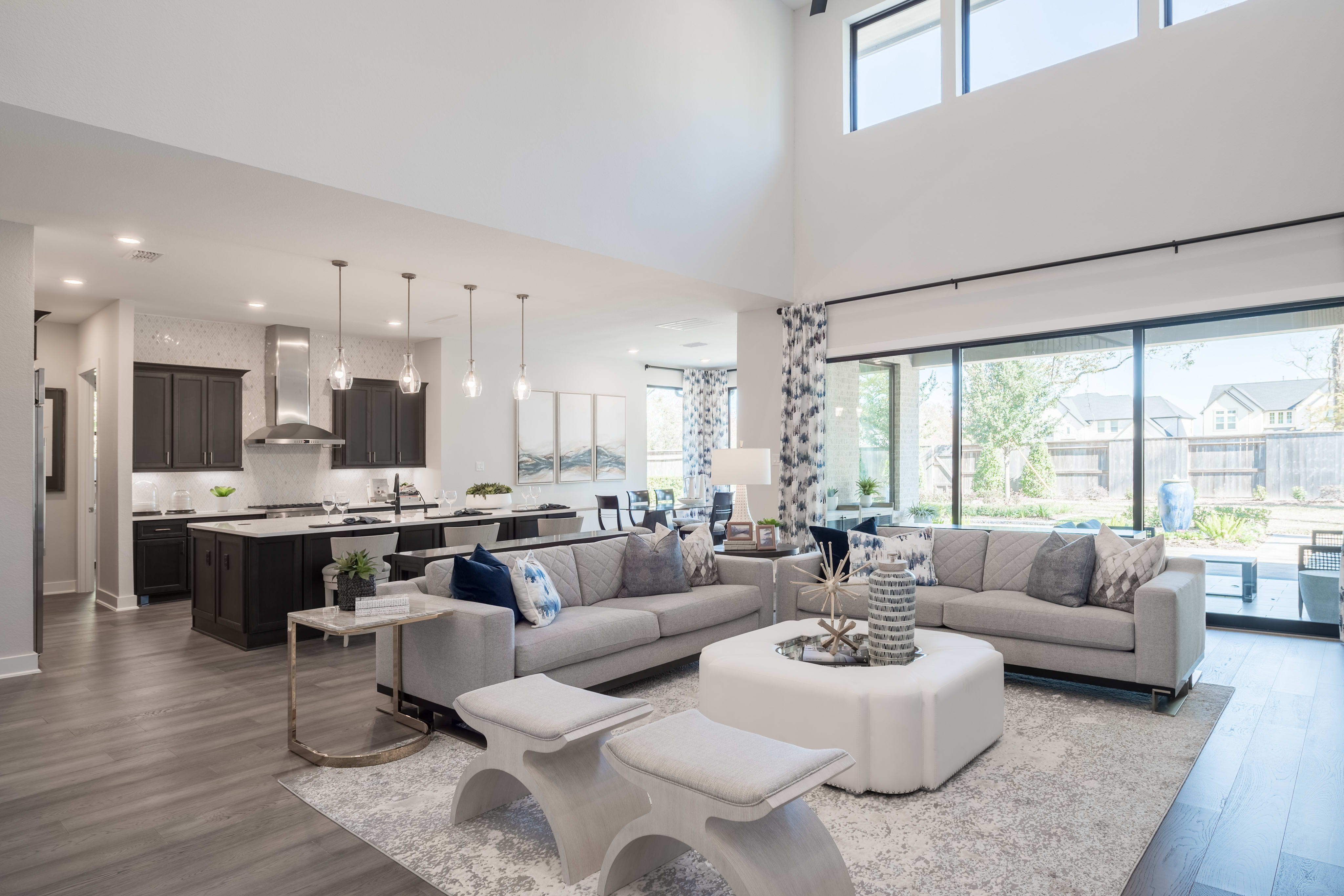 A modern and spacious open-concept living room with a kitchen visible in the background, featuring a gray sectional sofa, a round ottoman, and a large window providing natural light.