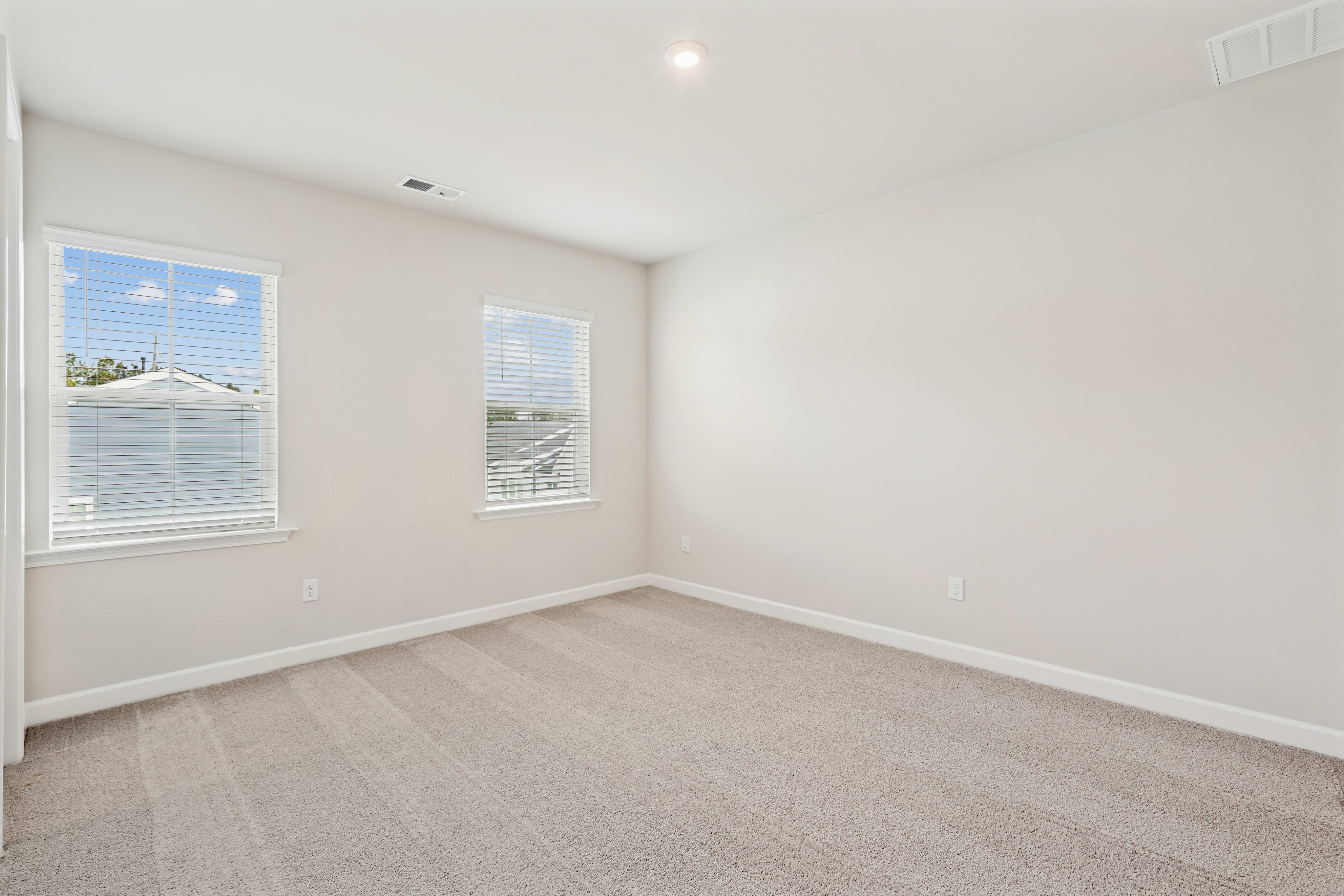 A spacious, well-lit room with white walls, gray carpeting, and large windows providing a view of the exterior.