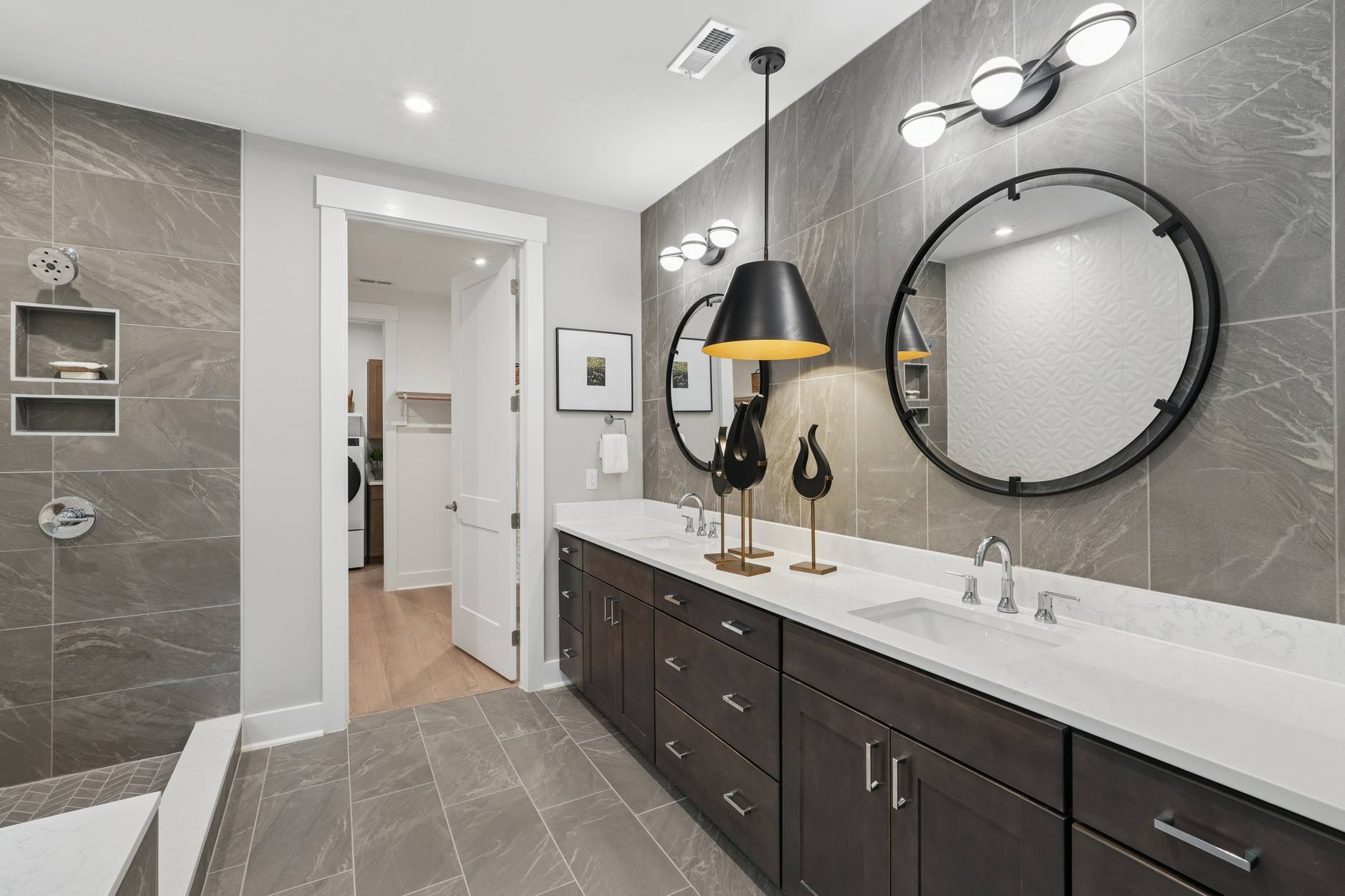 A modern and stylish bathroom with a large vanity, a round mirror, and various lighting fixtures, set against a backdrop of gray tiled walls.