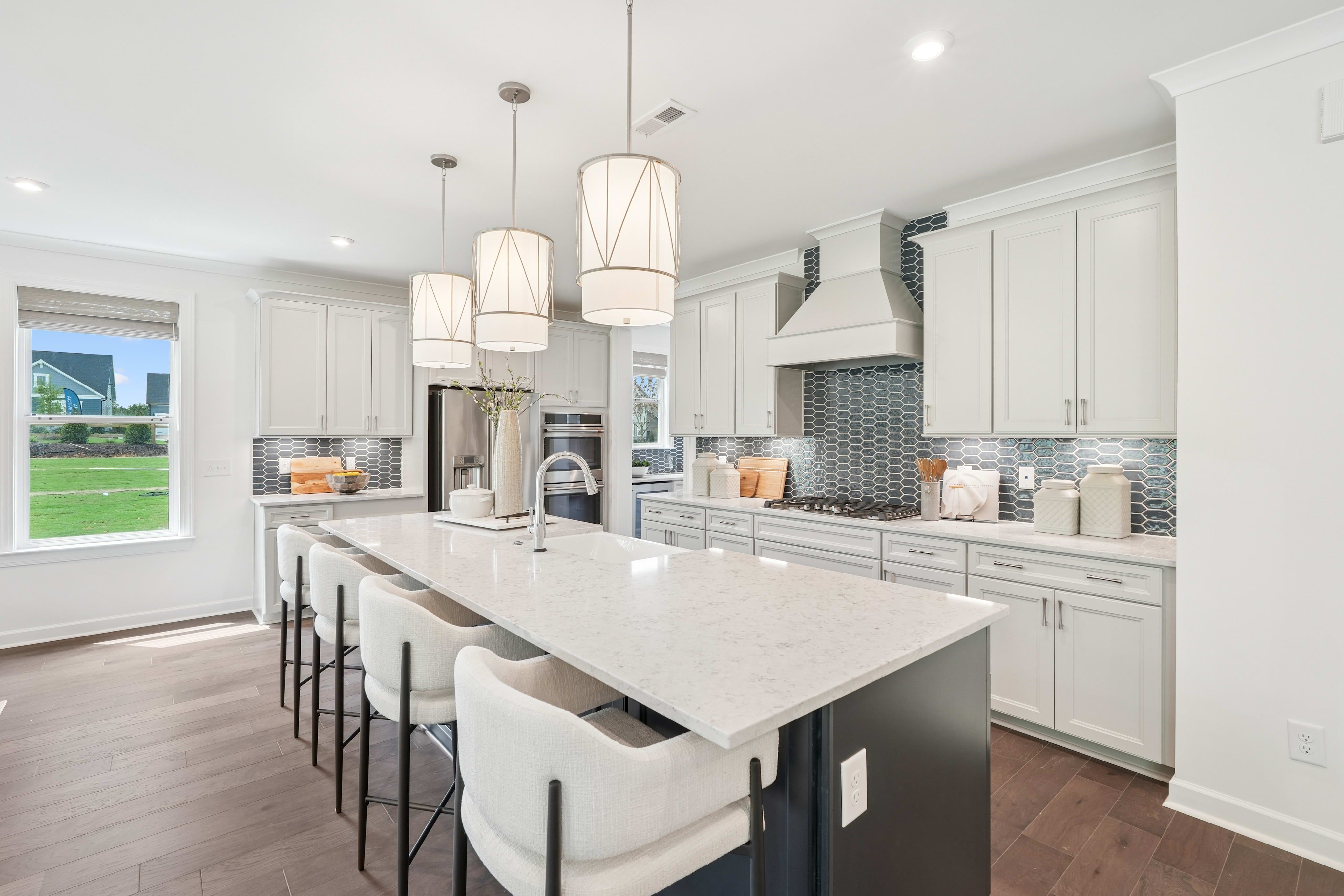 A modern, well-lit kitchen with white cabinets, a large island with bar stools, and pendant lighting fixtures hanging above.
