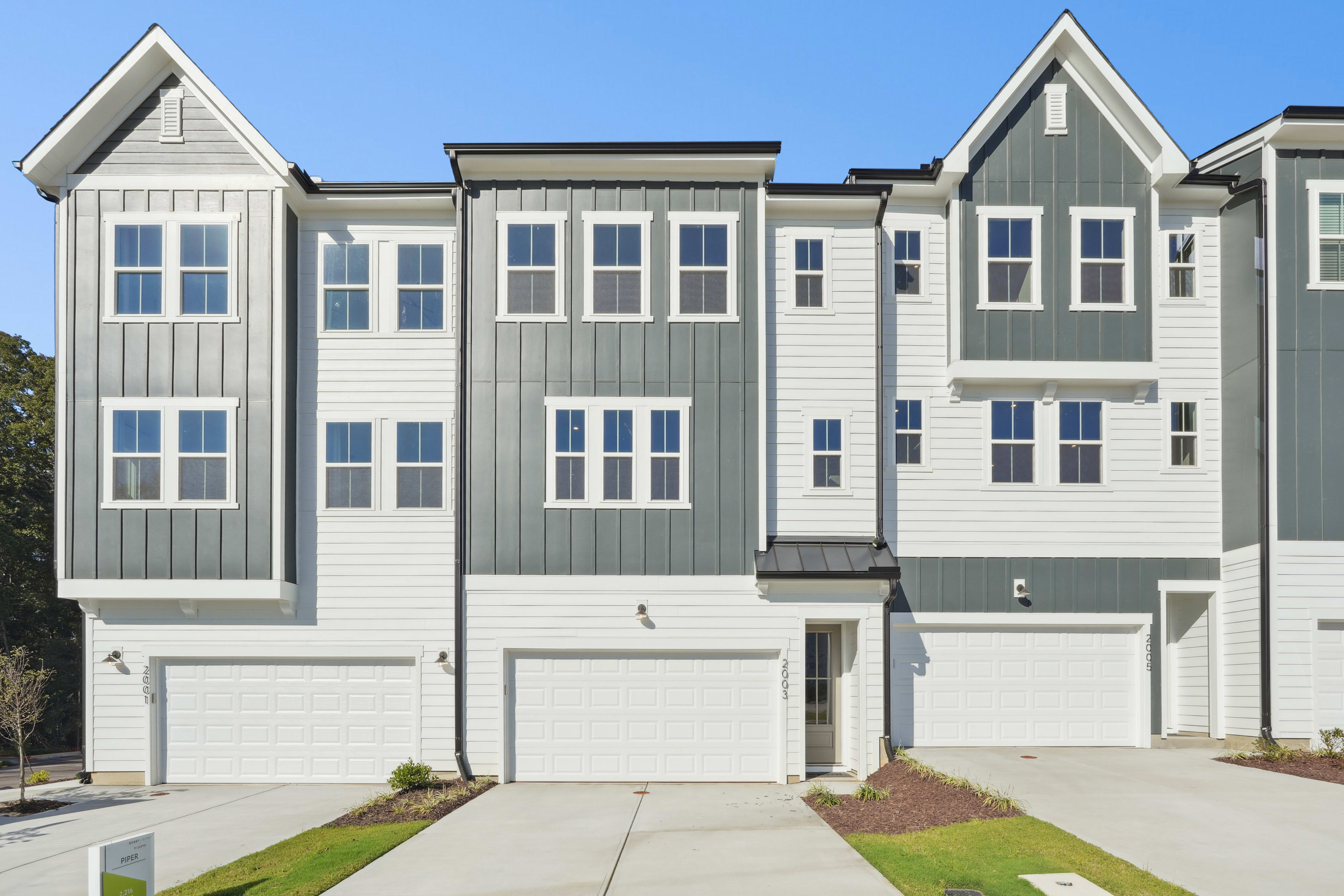 A row of modern, multi-story townhouses with gray and white siding, gabled roofs, and attached garages, set against a clear blue sky.