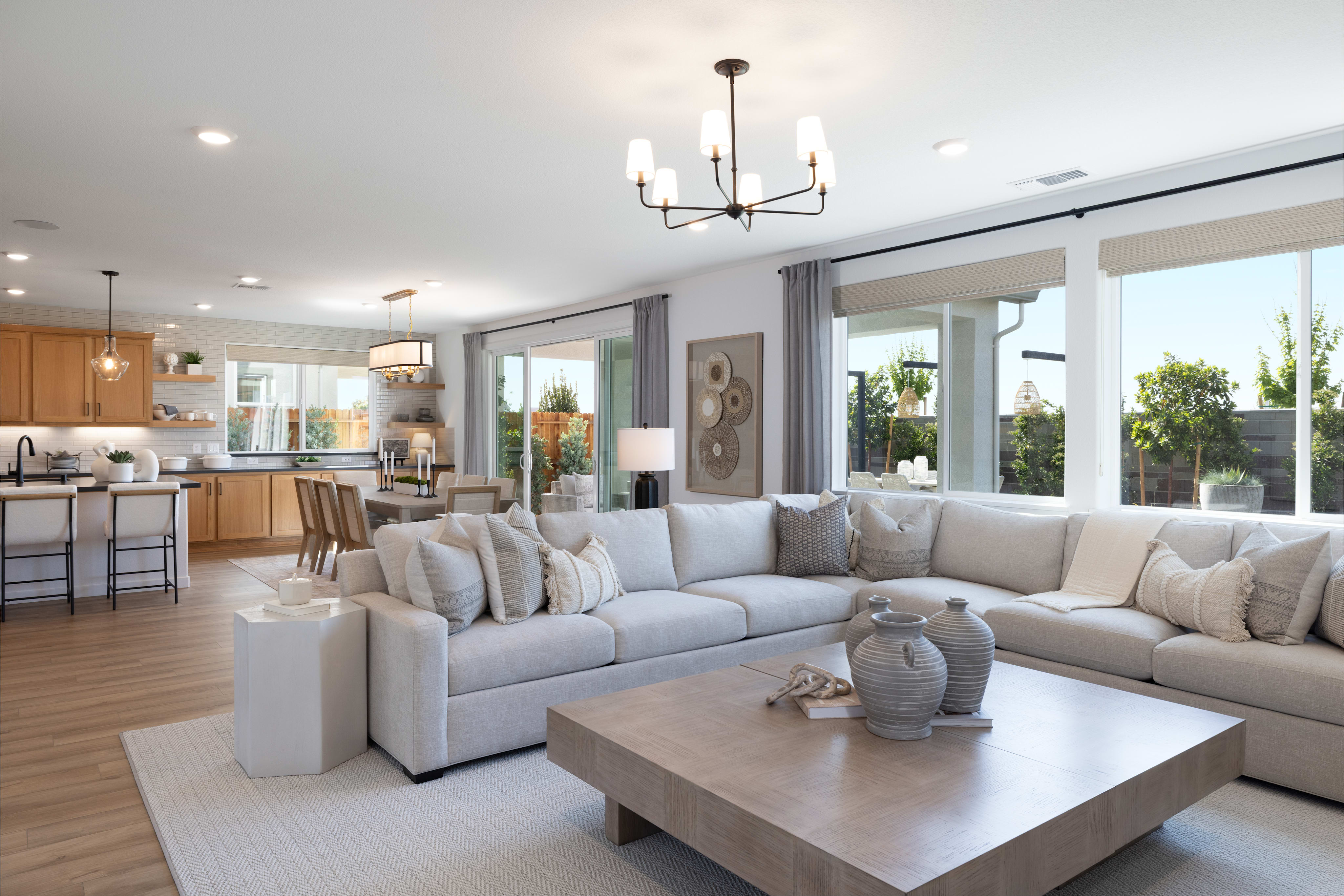 A spacious and modern living room with a large sectional sofa, a wooden coffee table, and a chandelier hanging from the ceiling. The room has large windows that provide natural light and a view of the outdoor scenery.