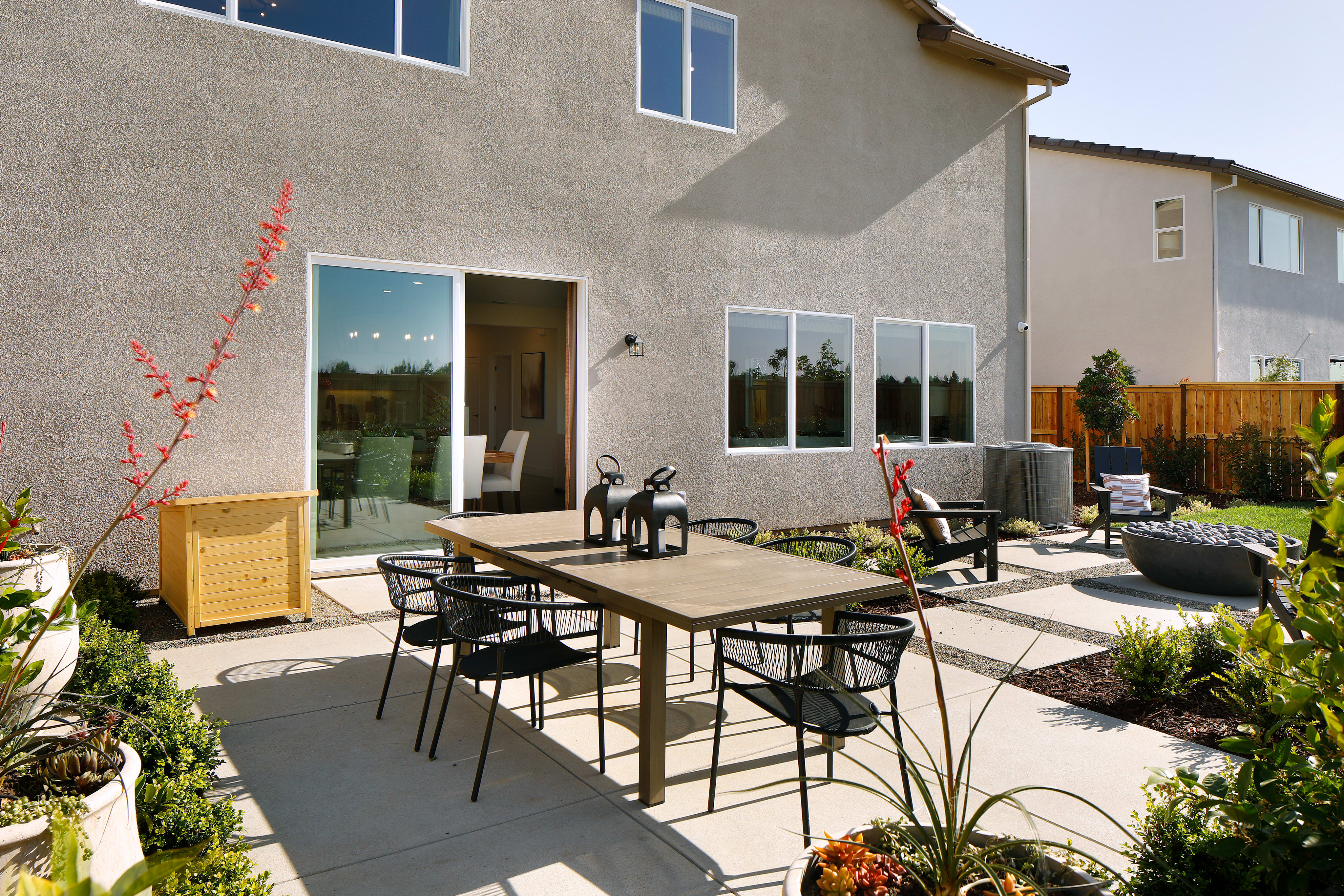 A well-designed outdoor patio area with a dining table and chairs, surrounded by lush greenery and a modern, stucco-clad home in the background.