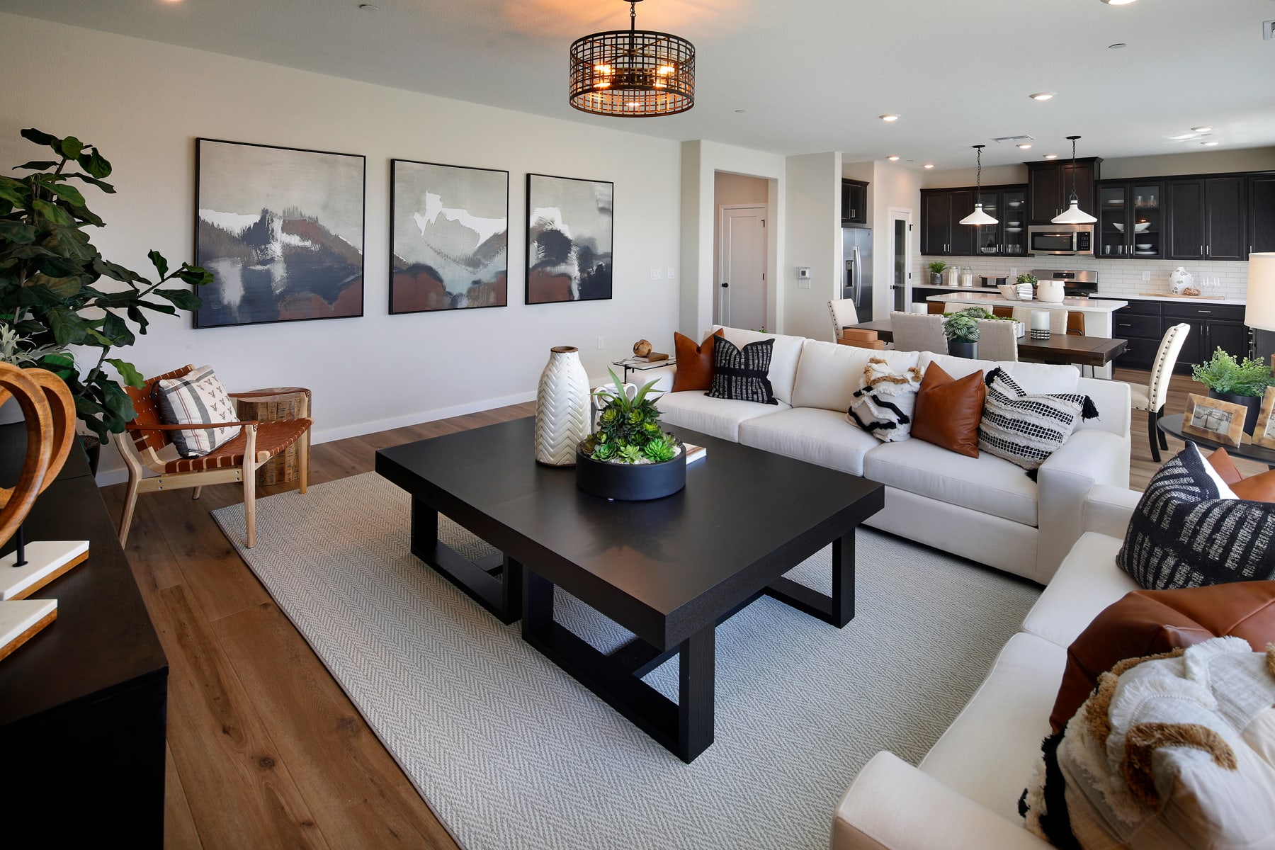 A cozy and modern living room with a large gray sofa, a black coffee table, and three framed landscape paintings on the wall. The kitchen can be seen in the background, with dark cabinets and a pendant light fixture.