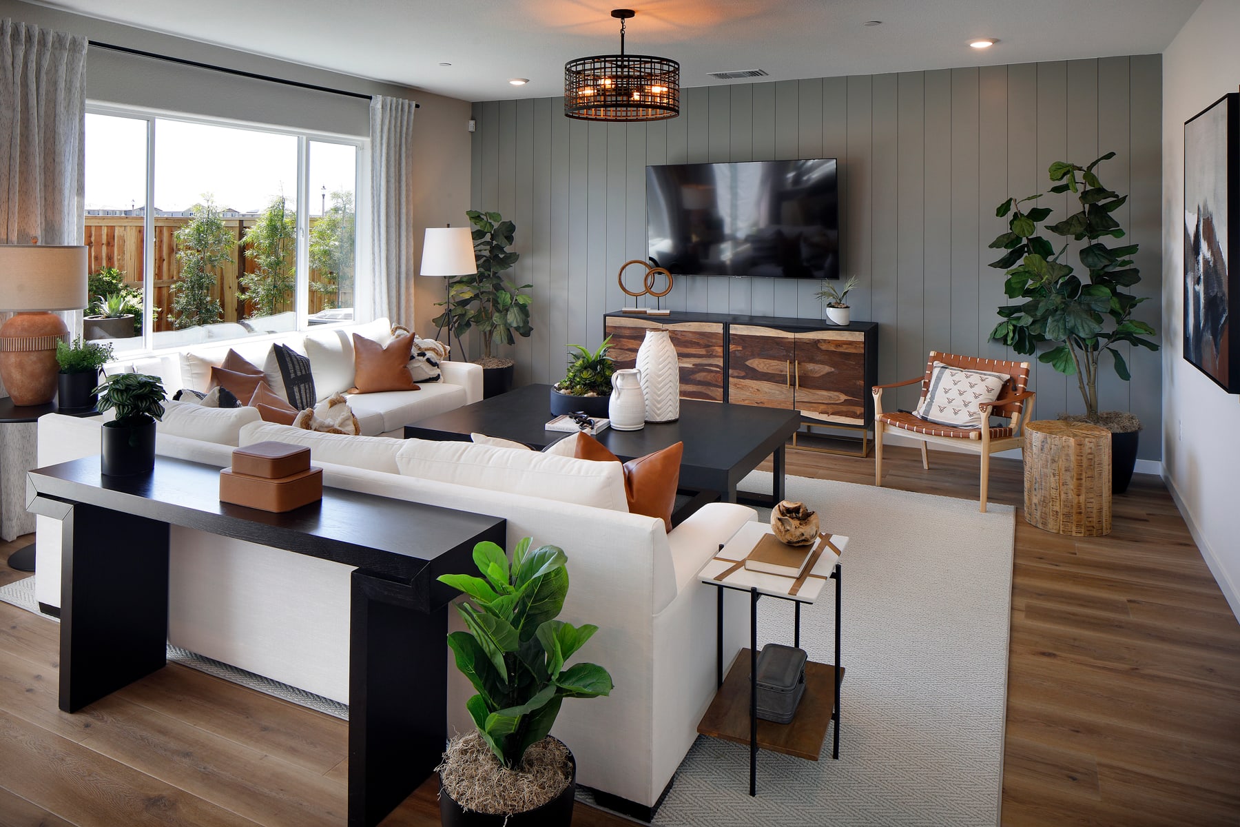 A cozy and well-decorated living room with a large TV, comfortable seating, and various decorative elements such as plants, artwork, and a stylish light fixture.