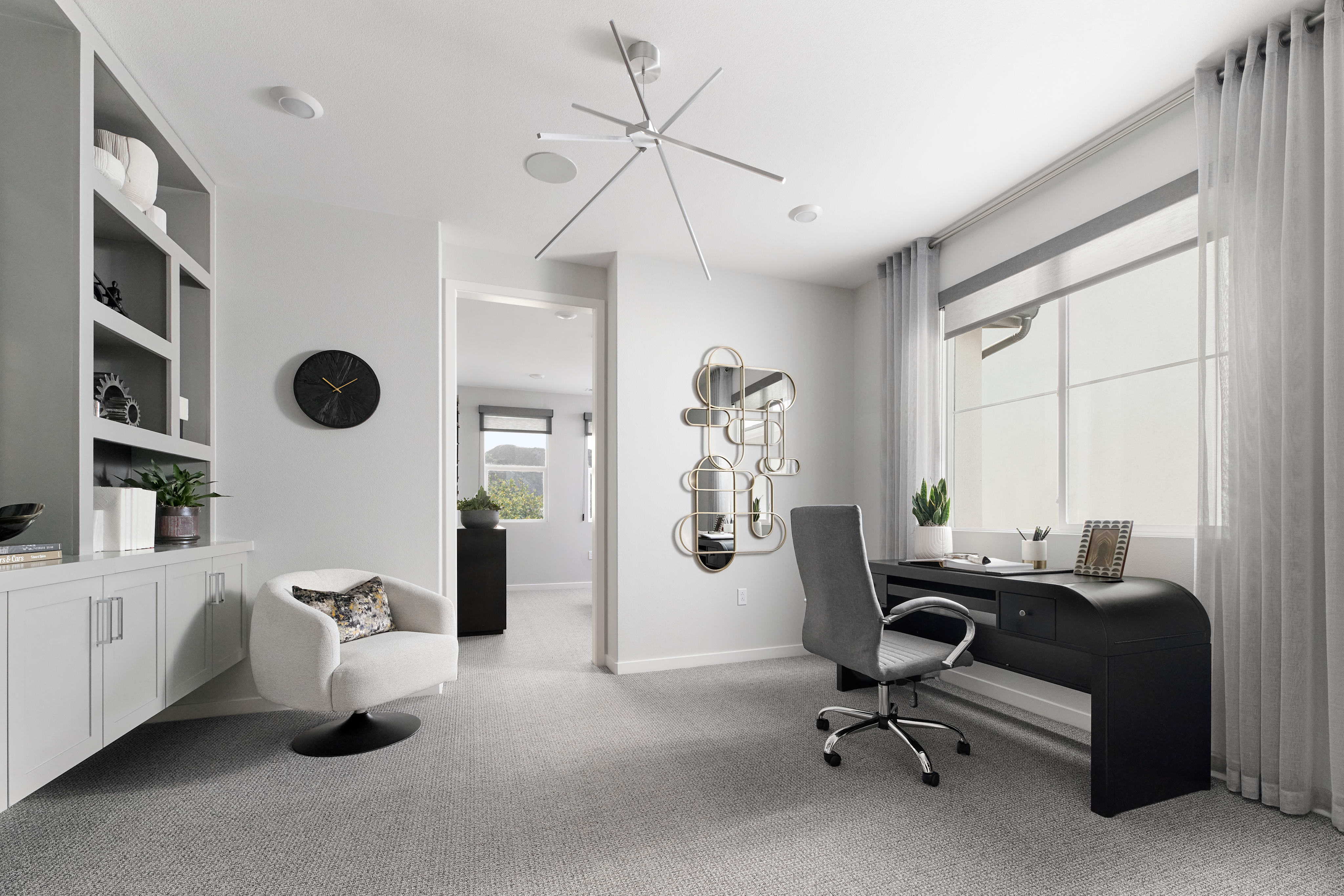 A modern, minimalist home office space with white walls, a black desk, and various decorative elements such as a ceiling fan, wall clocks, and potted plants.