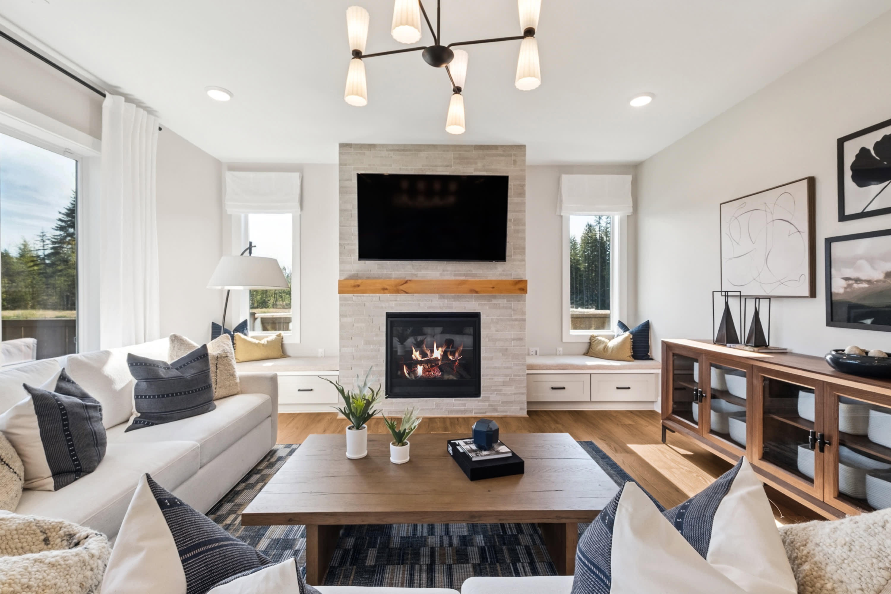 A cozy and inviting living room with a fireplace, wooden furniture, and a modern chandelier, surrounded by large windows that provide a scenic view of the outdoors.