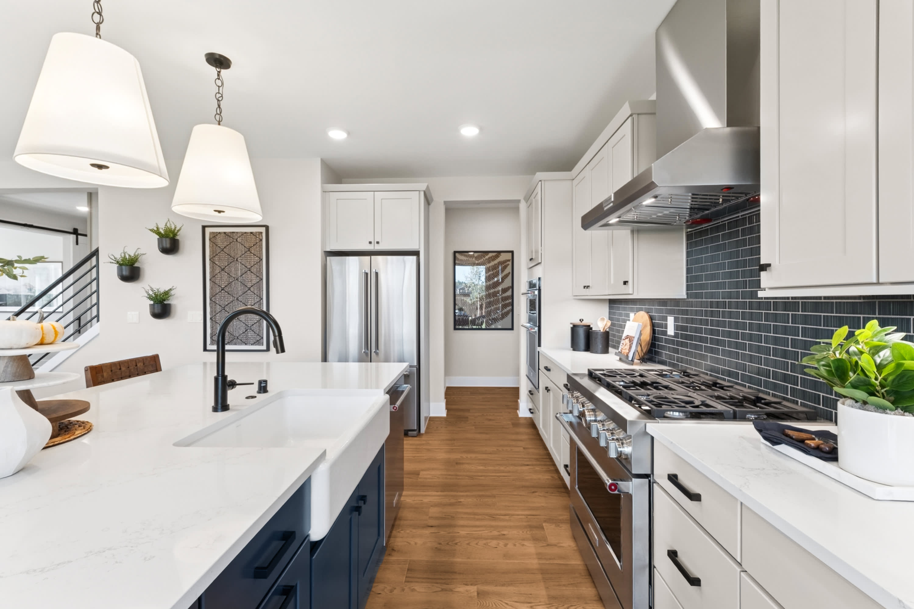 A modern, well-lit kitchen with white cabinets, a black tile backsplash, and stainless steel appliances, featuring a farmhouse-style sink and wooden flooring.