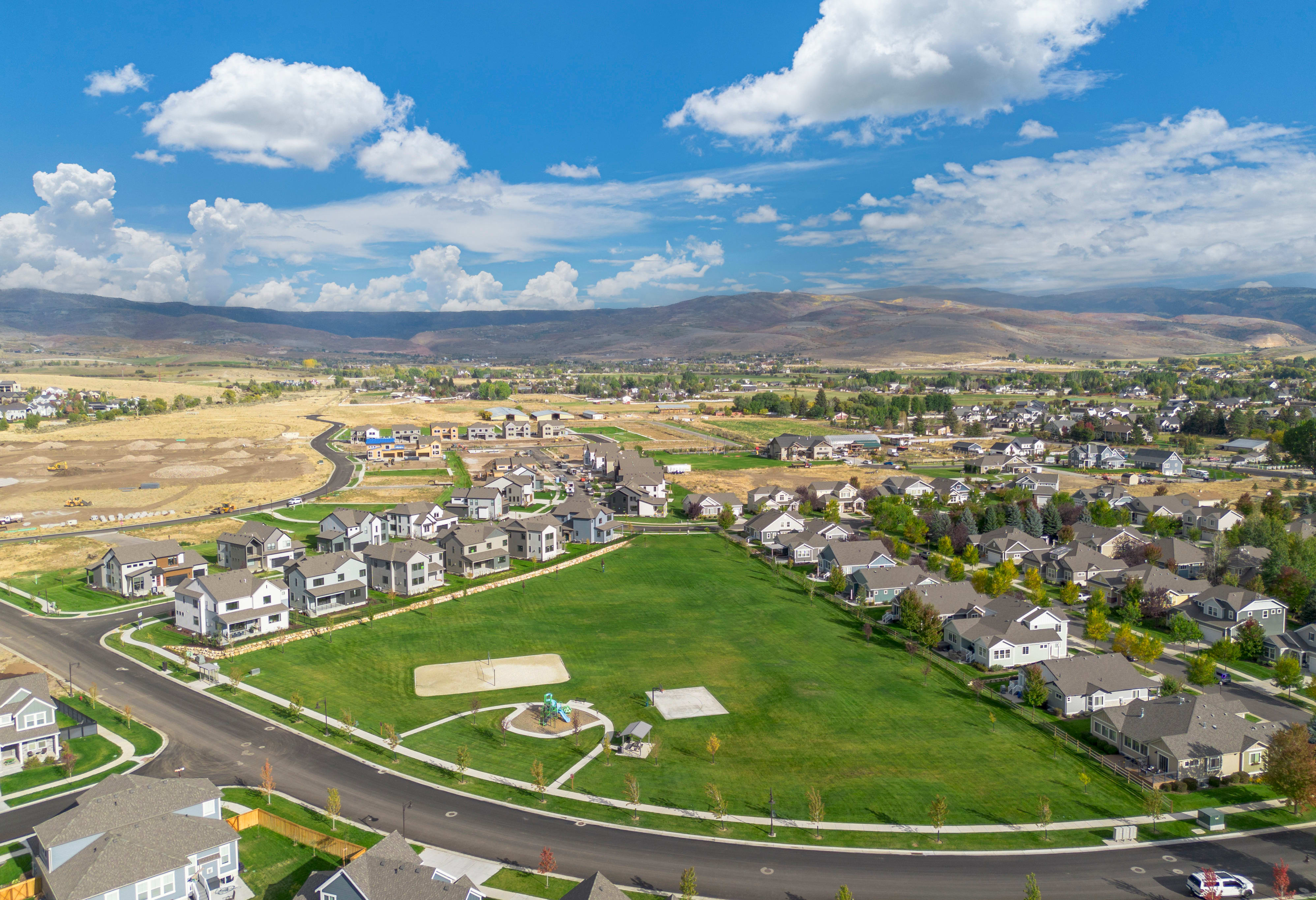 A sprawling suburban neighborhood nestled in a lush, rolling landscape, with a patchwork of green fields and winding roads leading up to the distant mountains under a picturesque cloudy sky.