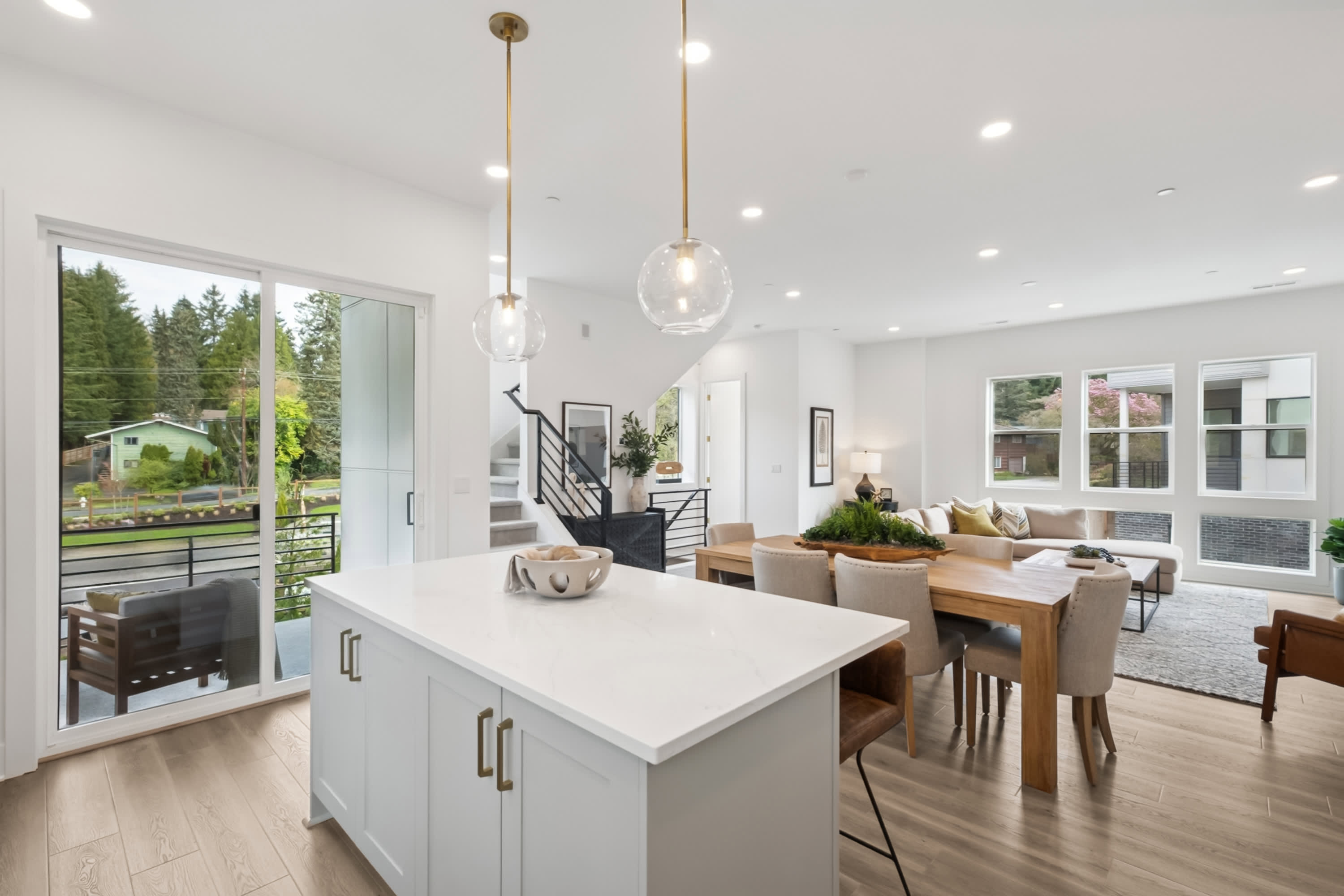 A modern, open-concept kitchen and living room with white walls, hardwood floors, and large windows overlooking a wooded outdoor area.