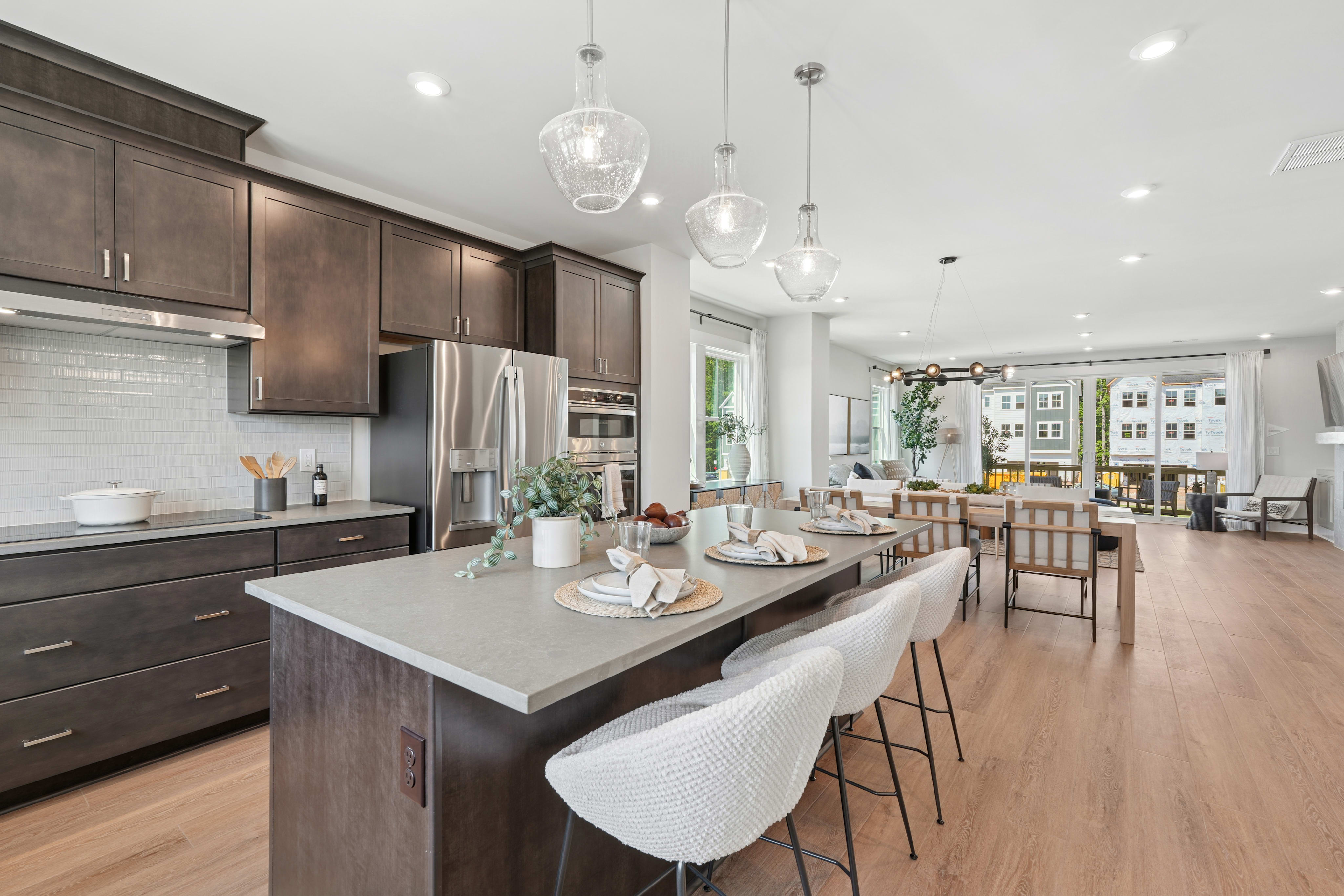 A modern and spacious kitchen with dark wood cabinets, a large island with a white countertop, and pendant lights hanging above, leading into an open living area with hardwood floors.