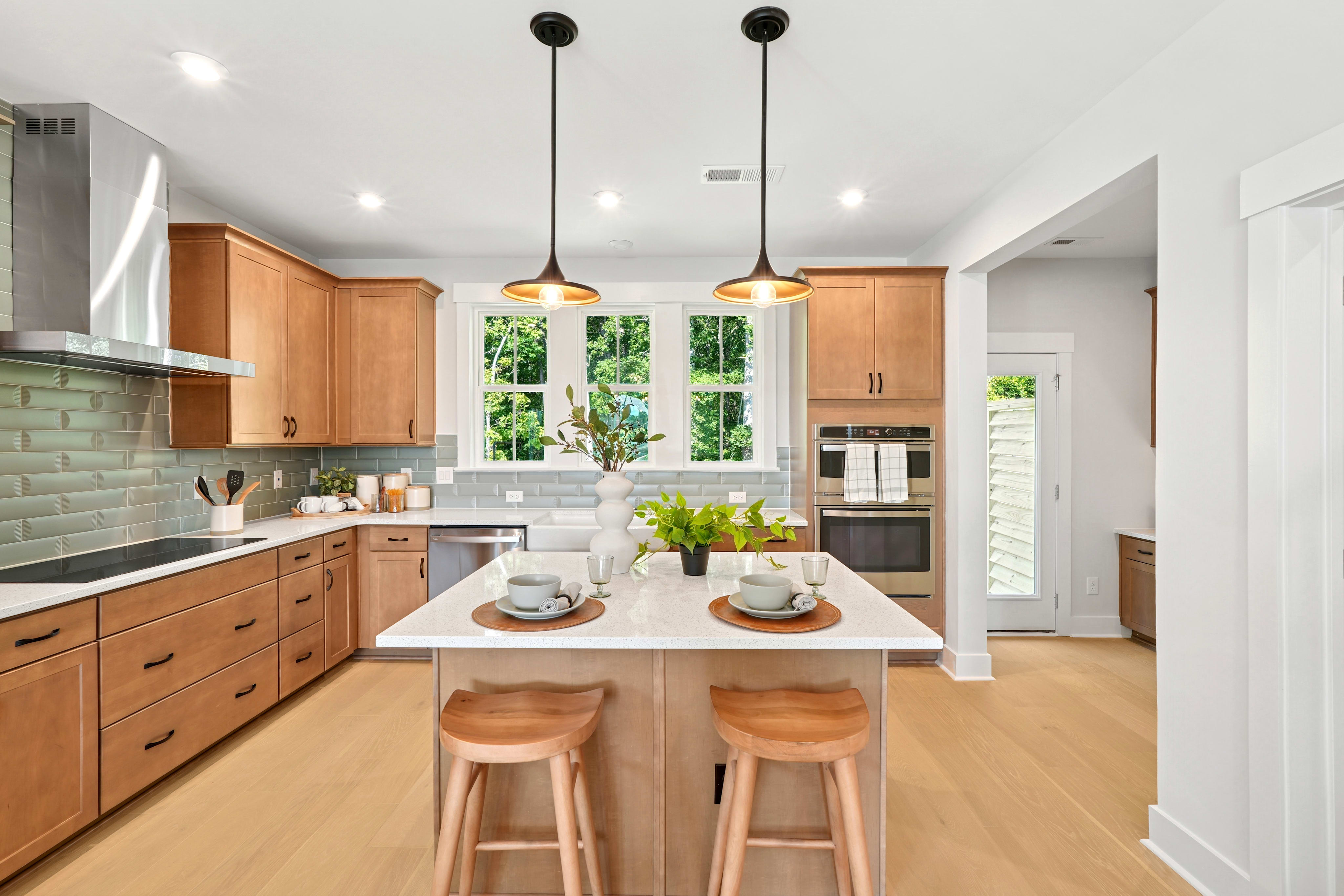 A modern, well-lit kitchen with wooden cabinets, a white countertop, and pendant lights, overlooking a lush outdoor view through large windows.