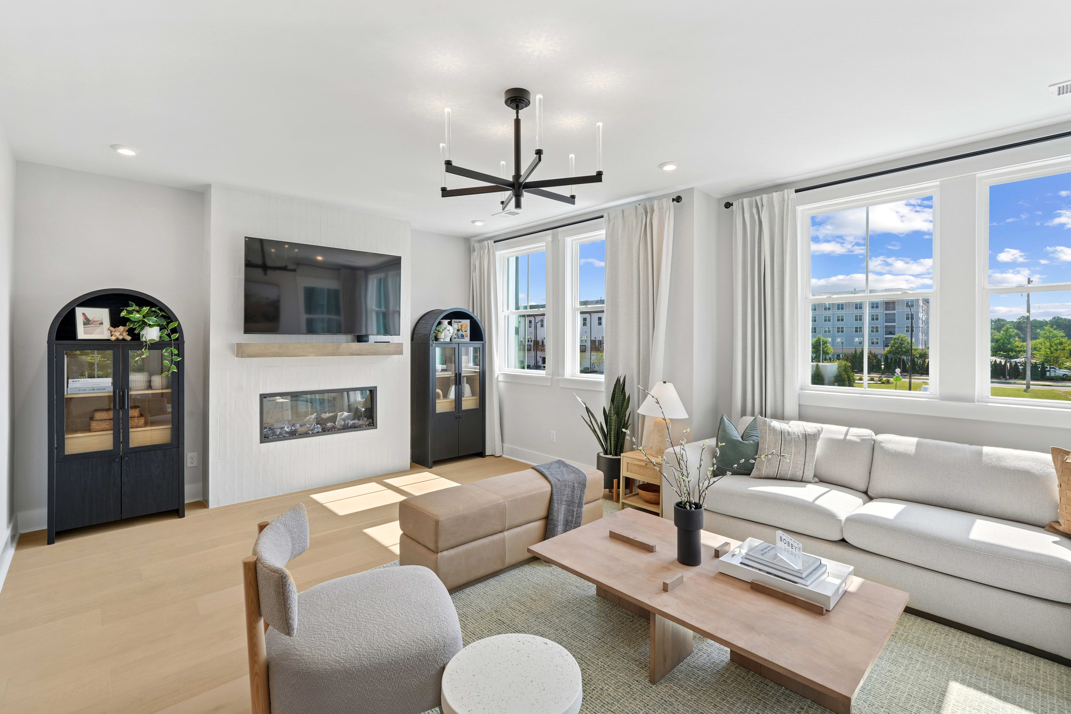 A modern and bright living room with a fireplace, large windows, and a stylish chandelier, surrounded by comfortable furniture and decor.