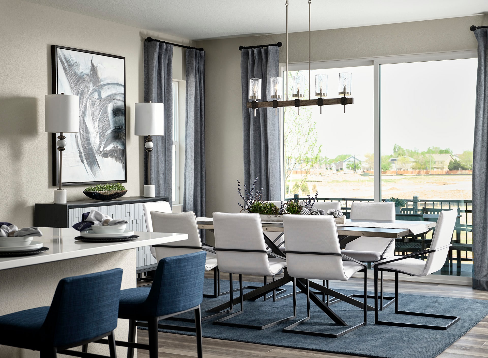 A modern and stylish dining room with a large window overlooking a scenic outdoor area, featuring a rectangular dining table surrounded by white and blue upholstered chairs, a black and white abstract artwork on the wall, and a sleek chandelier overhead.