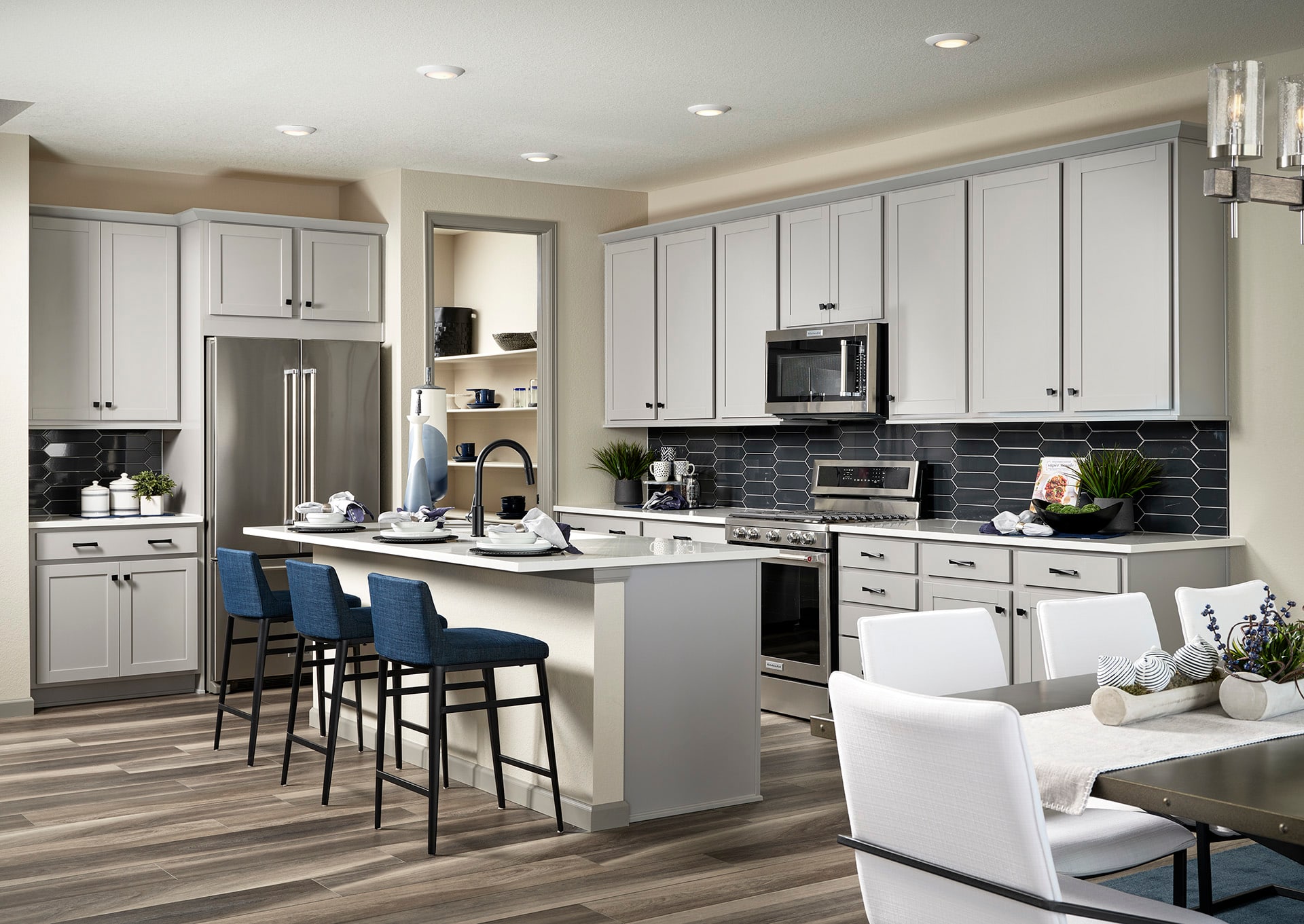 A modern, open-concept kitchen with white cabinets, a gray tile backsplash, and a central island with bar stools, all set against a backdrop of a cozy living area with a white sofa.