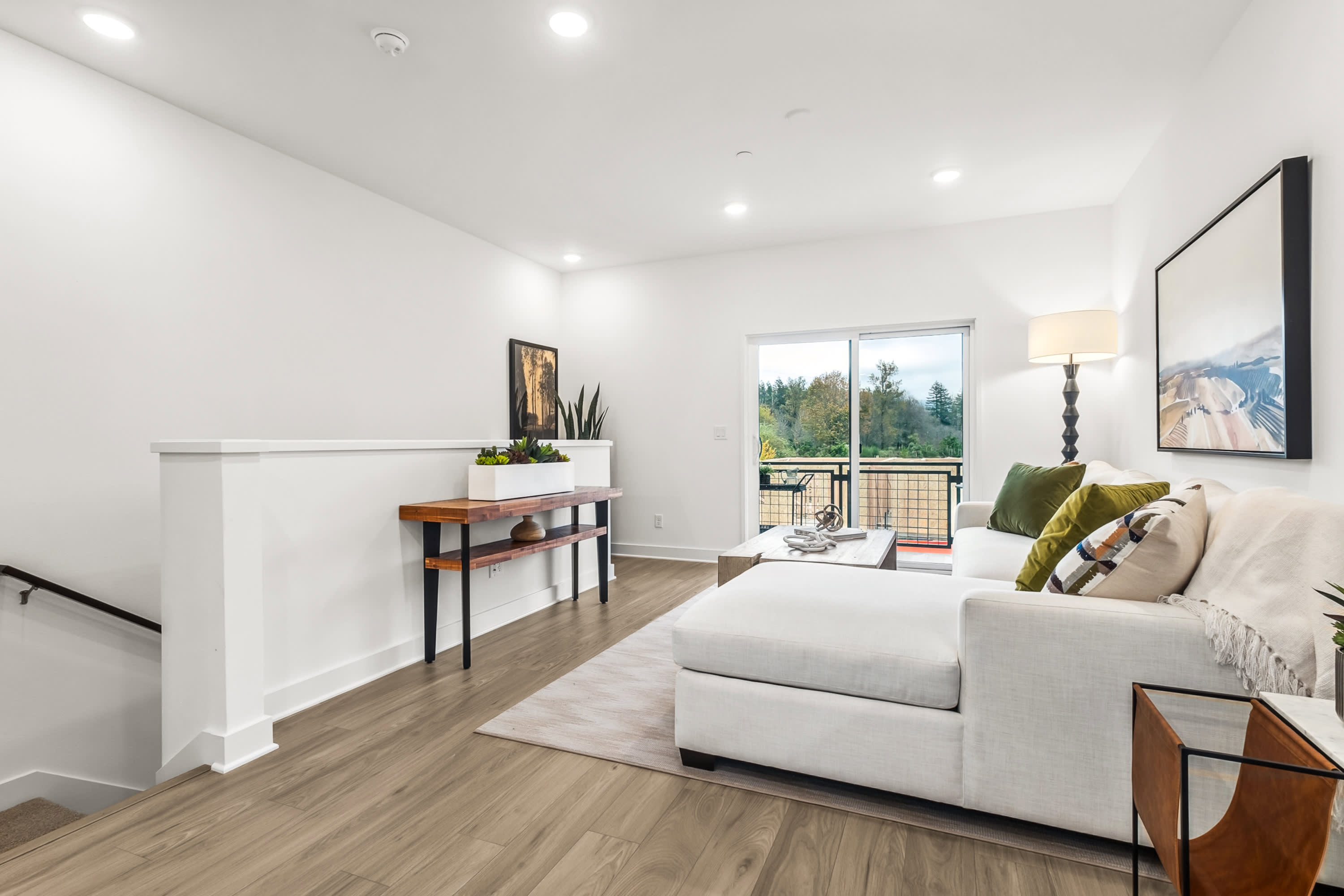 A modern and minimalist living room with a white couch, wooden console table, and a large window overlooking a forested landscape.