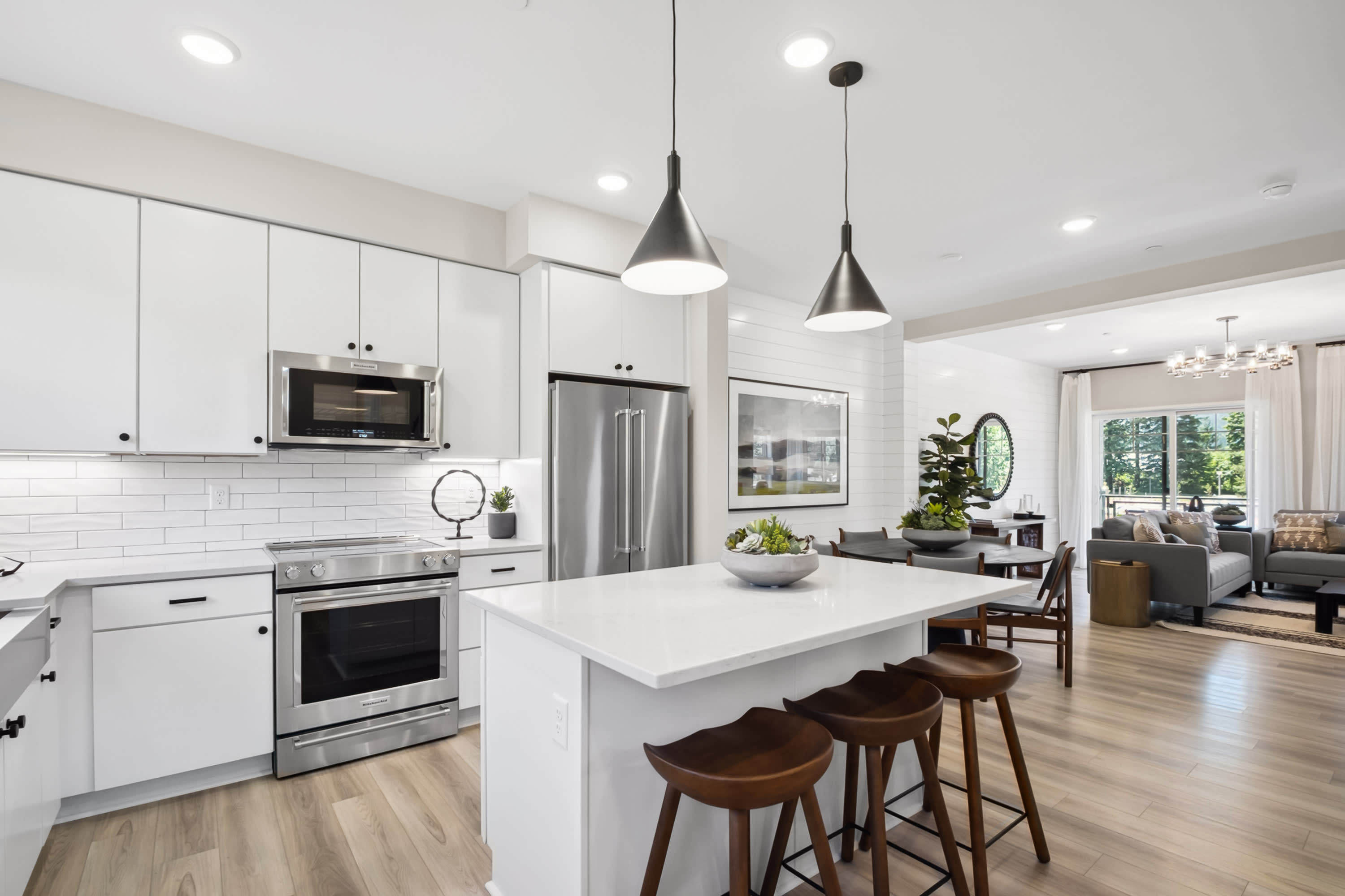 A modern, open-concept kitchen with white cabinets, stainless steel appliances, and a central island with wooden stools, leading into a cozy living room area with a sofa and armchair.