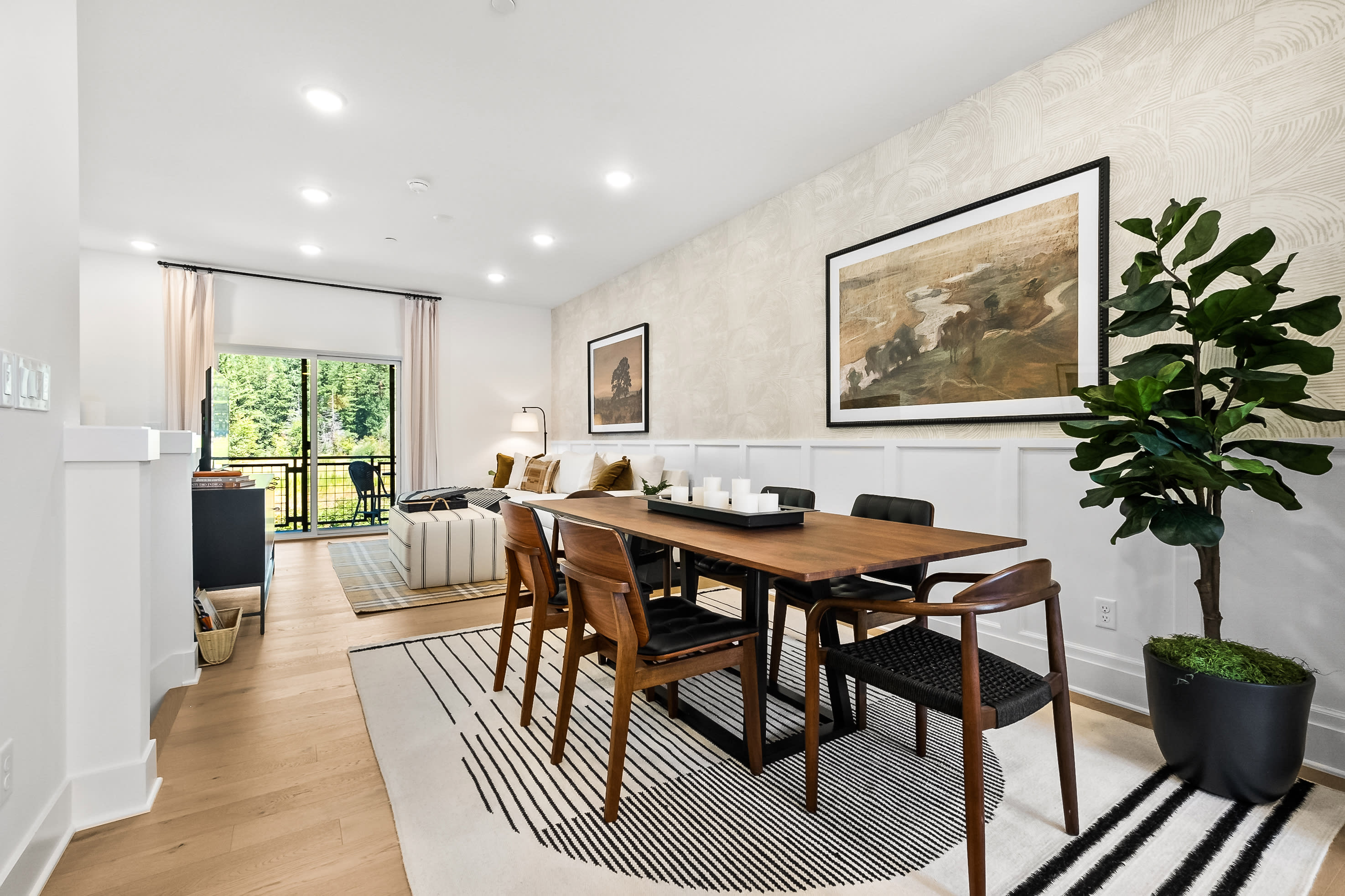 A modern and minimalist dining room with a wooden table, chairs, and a large framed artwork on the wall, surrounded by potted plants and natural lighting from the window.