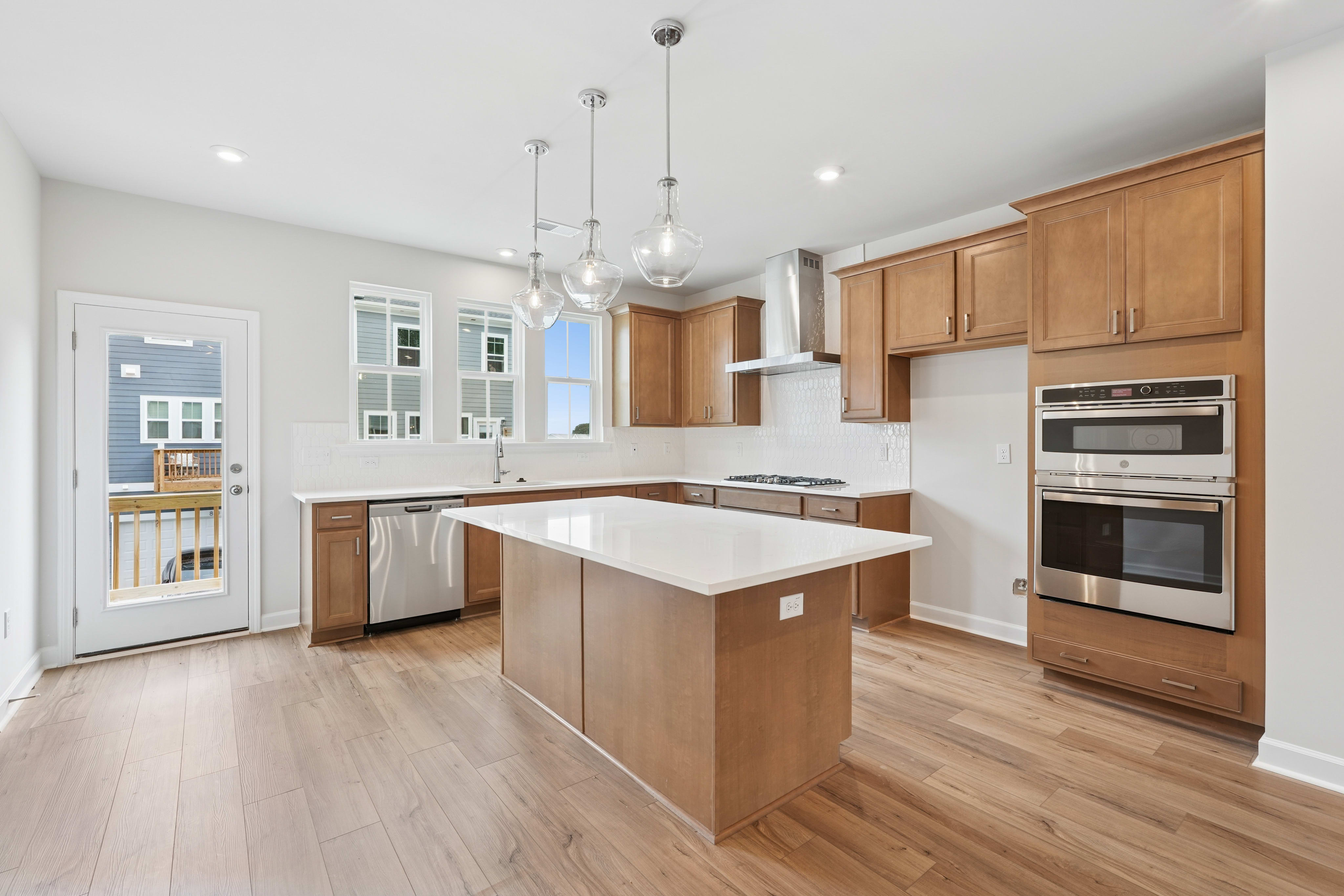A modern, open-concept kitchen with light wood cabinetry, a central island, and stainless steel appliances, set against a backdrop of white walls and hardwood floors.