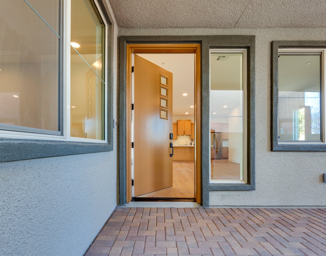 The image shows a modern entryway with a wooden door, glass panels, and a brick floor leading into a brightly lit interior.