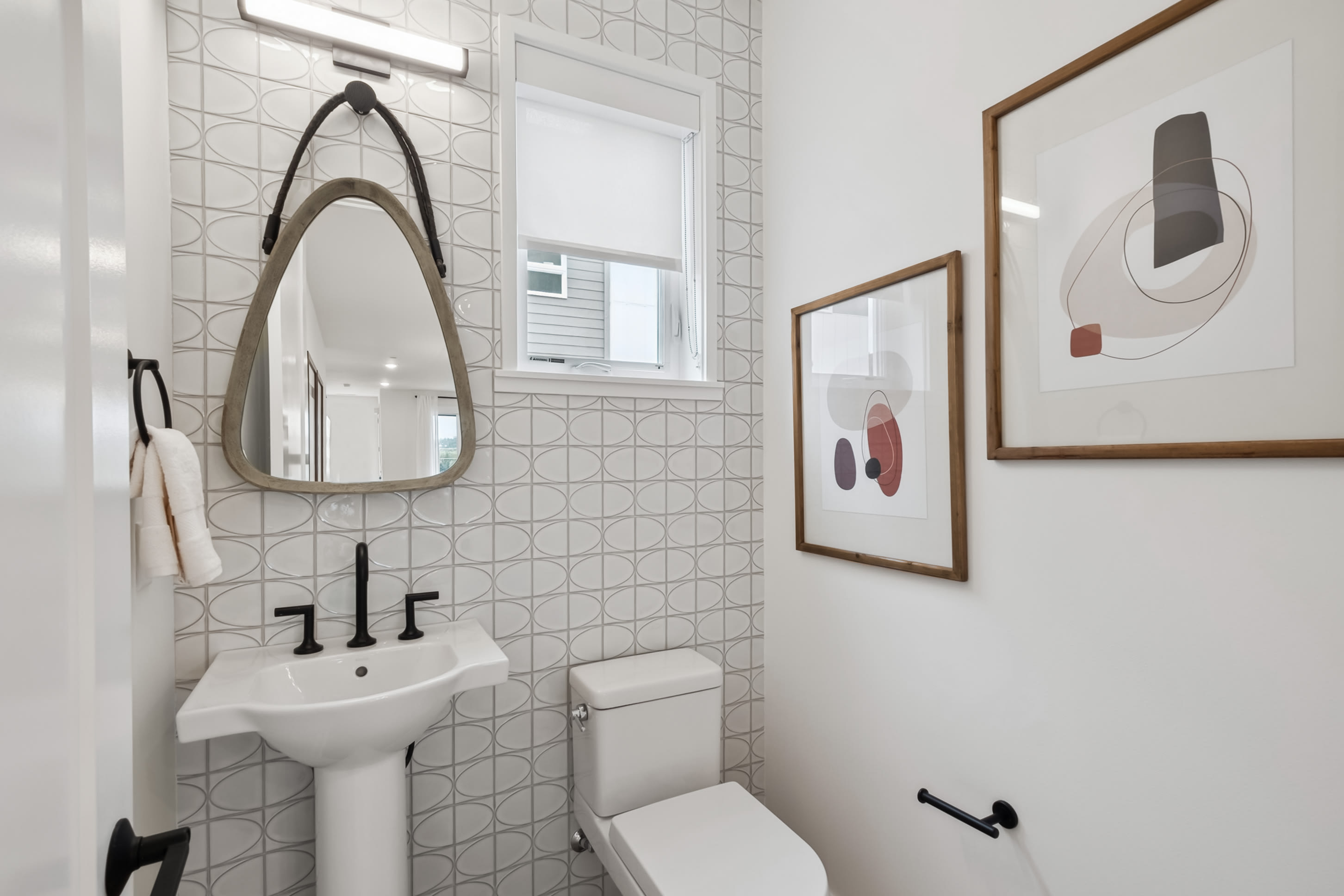 A modern, minimalist bathroom with white tile walls, a pedestal sink, and framed artwork on the walls.
