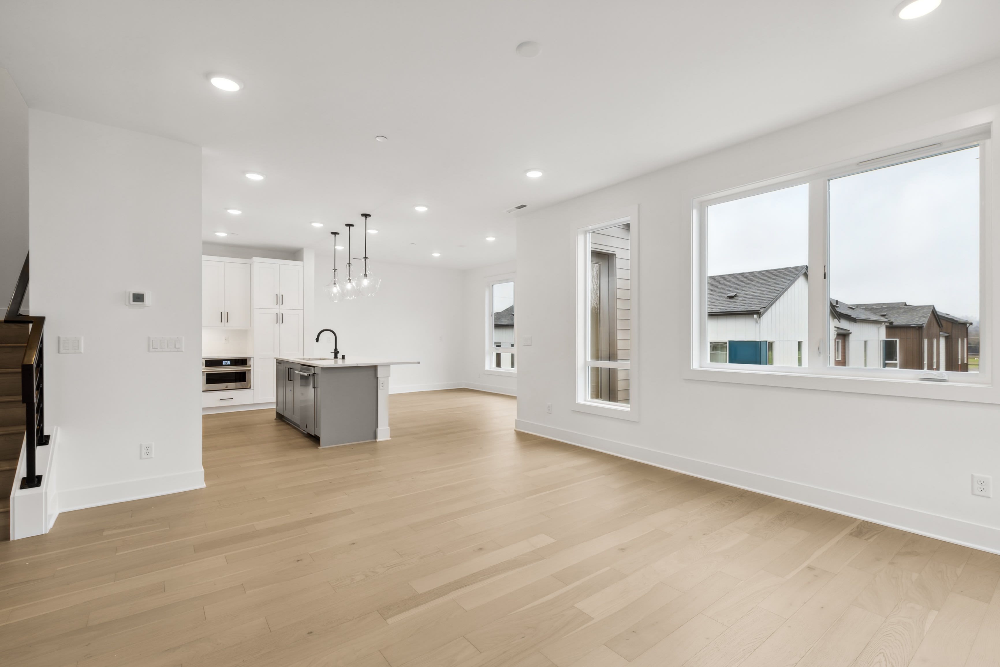 A spacious, modern kitchen with white walls, hardwood floors, and large windows overlooking a residential neighborhood in the background.