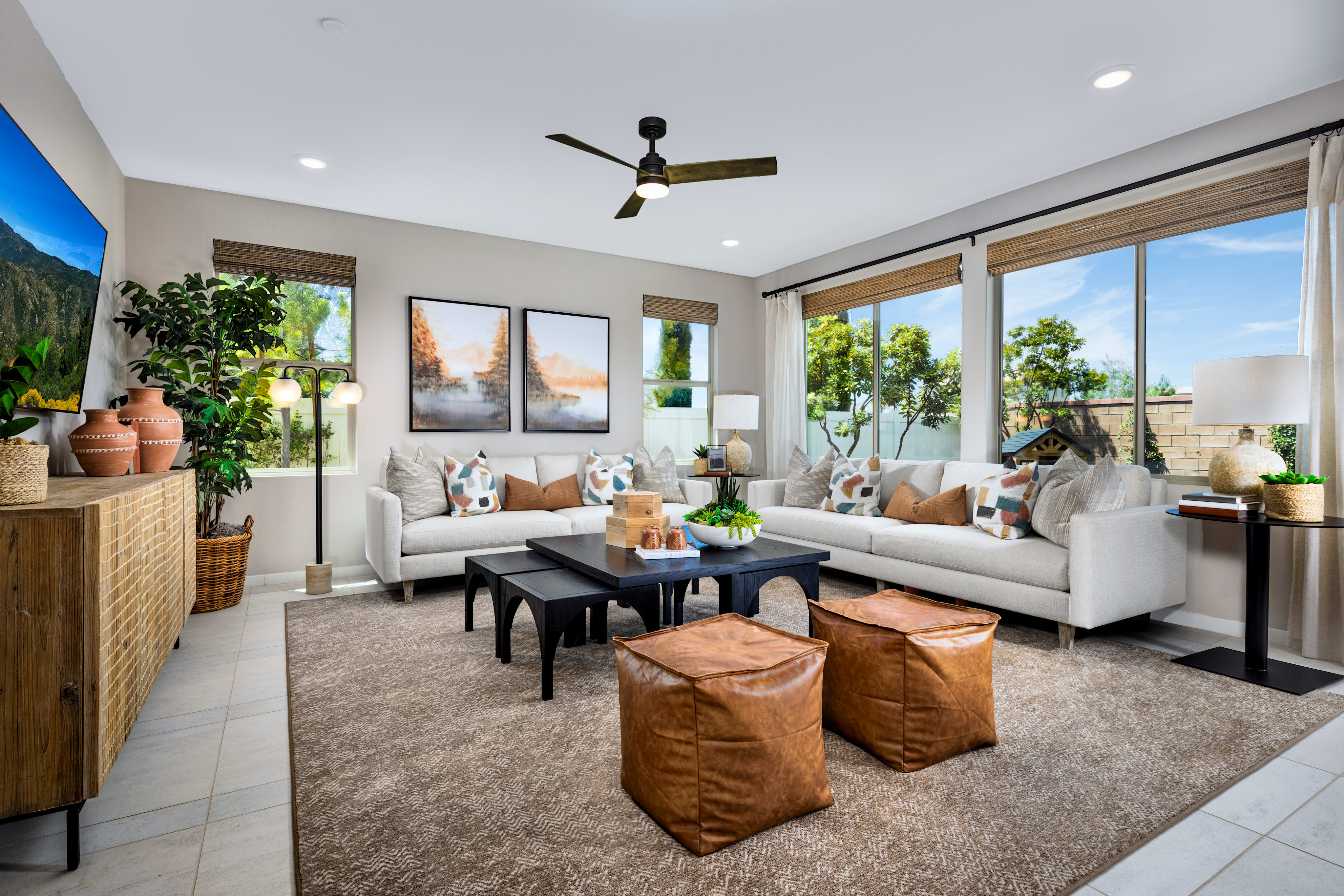 A modern and cozy living room with large windows, a ceiling fan, and various decorative elements such as plants, artwork, and leather ottomans.