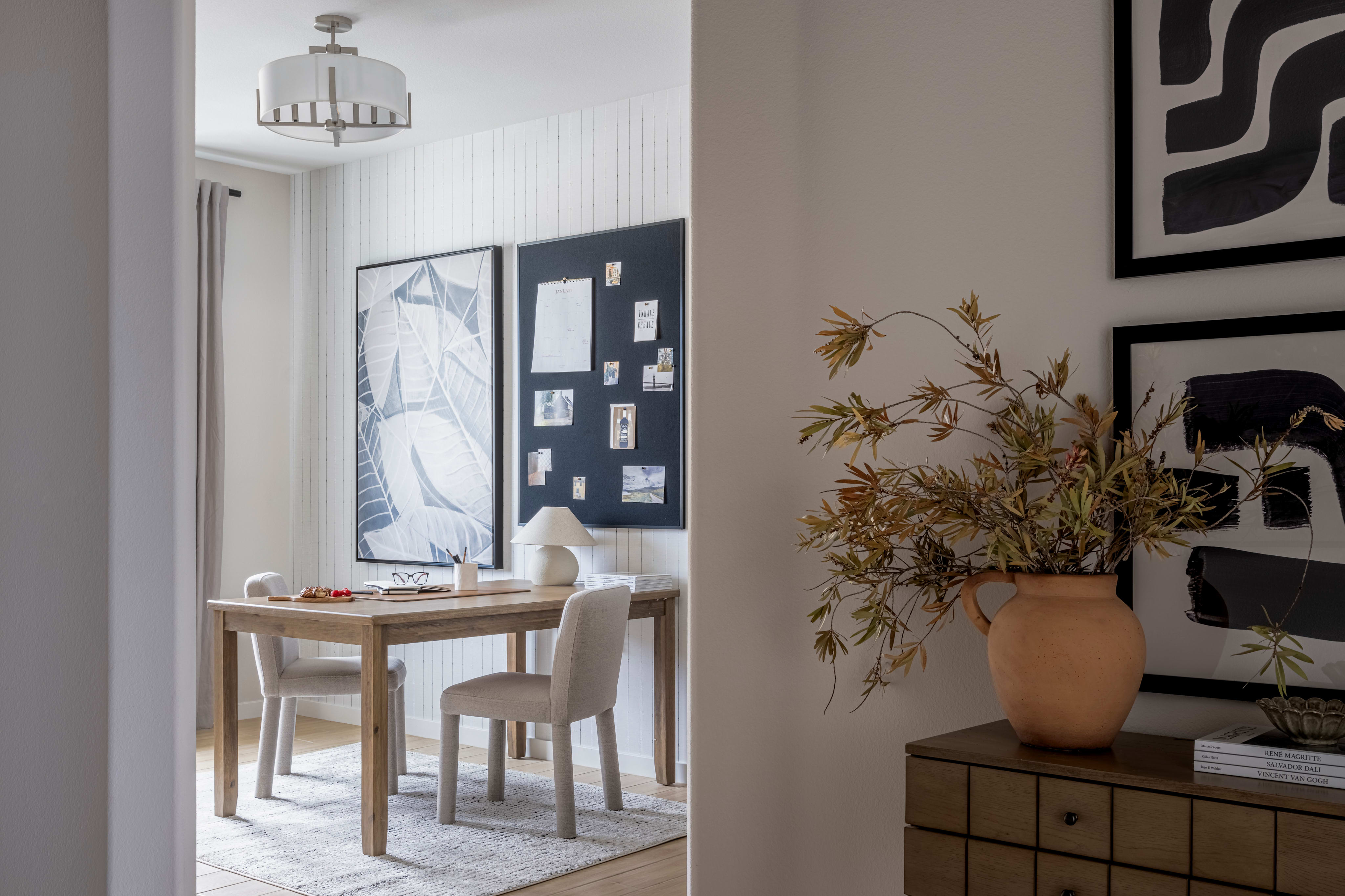 A cozy and well-designed living space with a wooden table, chairs, and a decorative vase in the foreground, leading to a hallway with artwork and a cabinet in the background.
