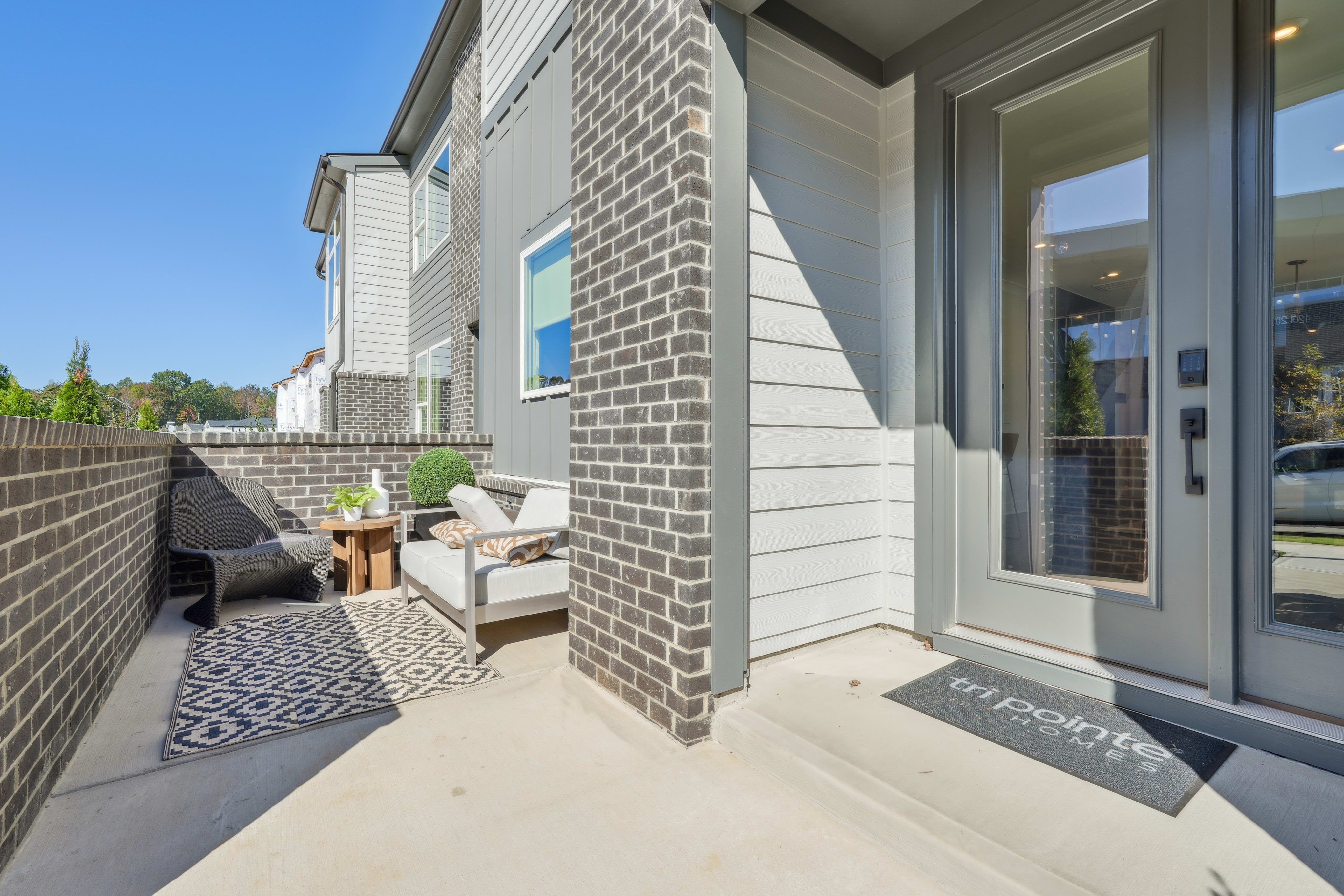 A cozy outdoor patio area with a brick wall, comfortable seating, and a decorative rug on the concrete floor, set against a backdrop of a residential building.