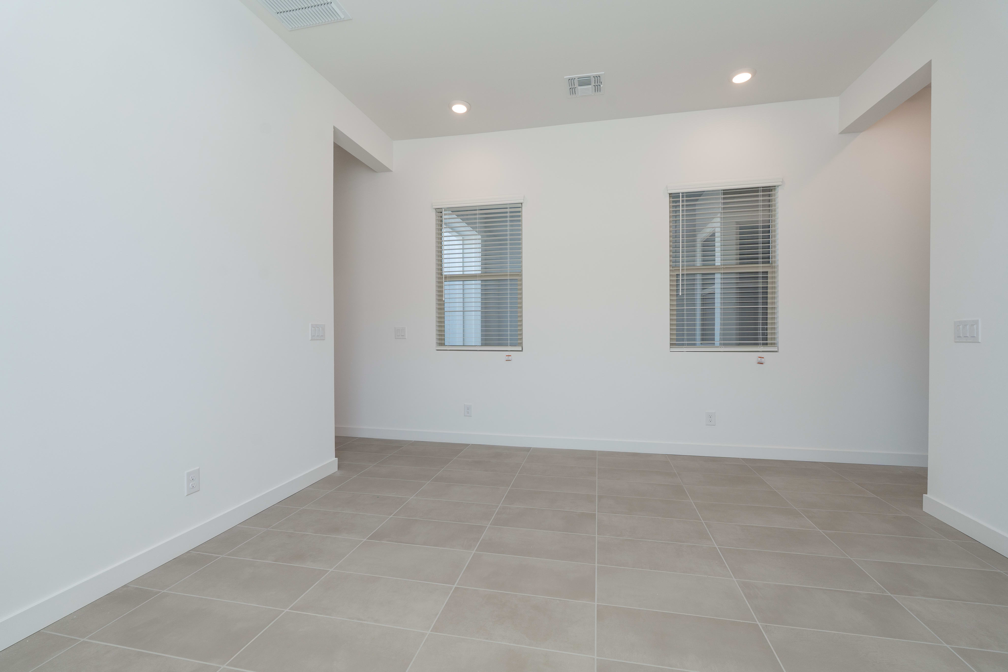 A spacious, well-lit room with white walls, recessed lighting, and large windows with blinds, featuring a tiled floor in a neutral color scheme.