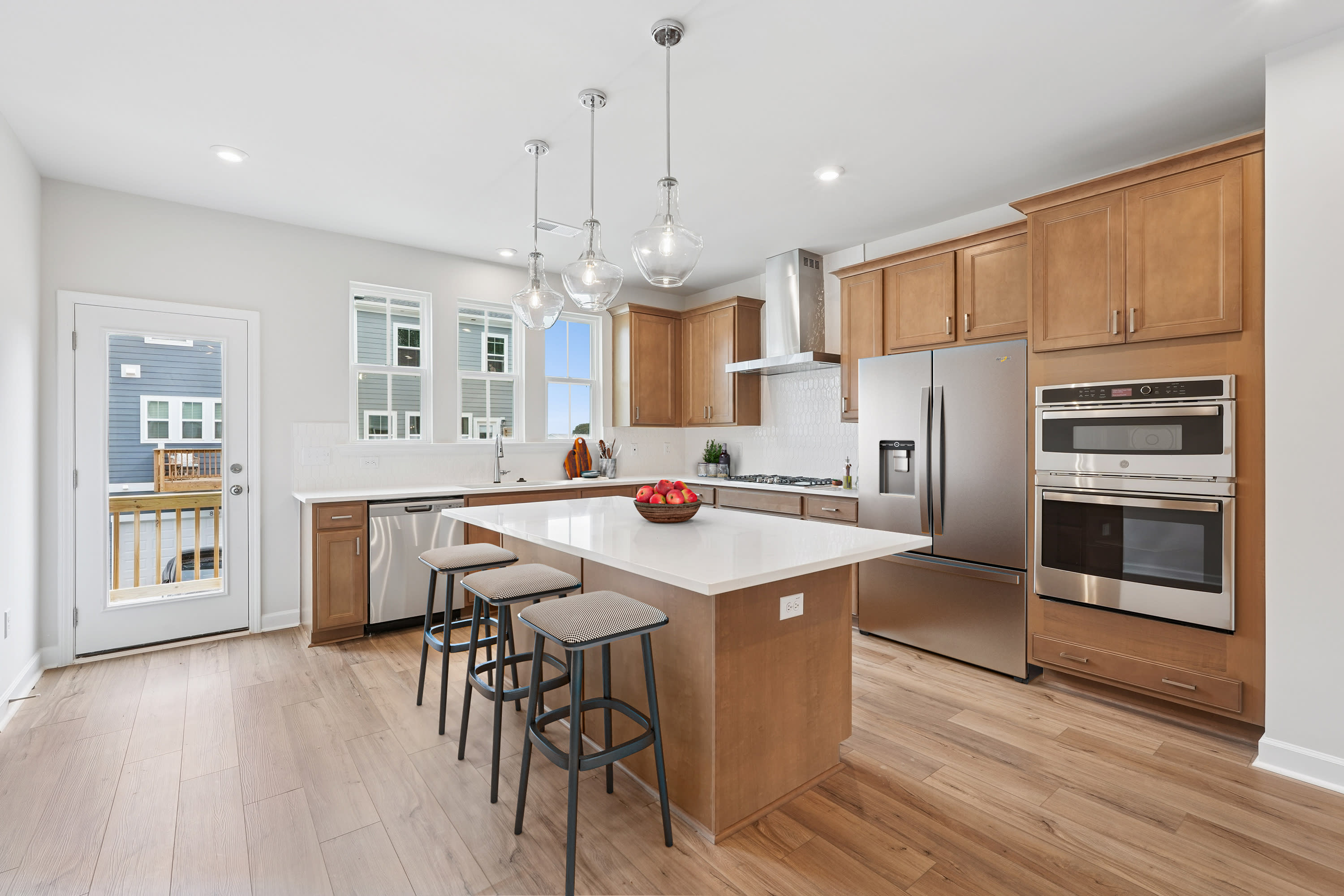 A modern, open-concept kitchen with wooden cabinets, stainless steel appliances, and a central island with bar stools, set against a backdrop of white walls and hardwood floors.