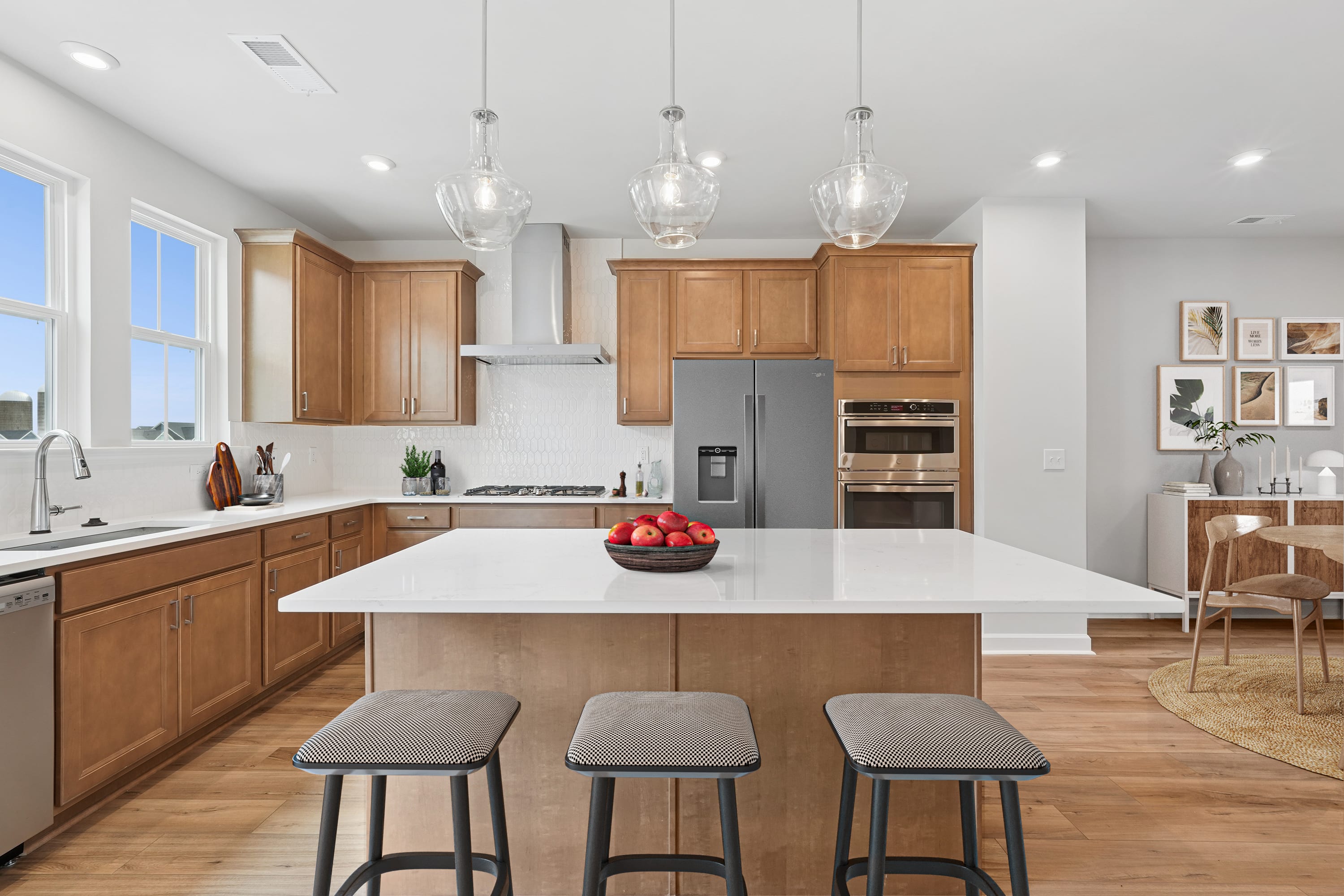 A modern, open-concept kitchen with wooden cabinets, a white countertop island, and pendant lighting fixtures, set against a backdrop of a bright, airy living space.