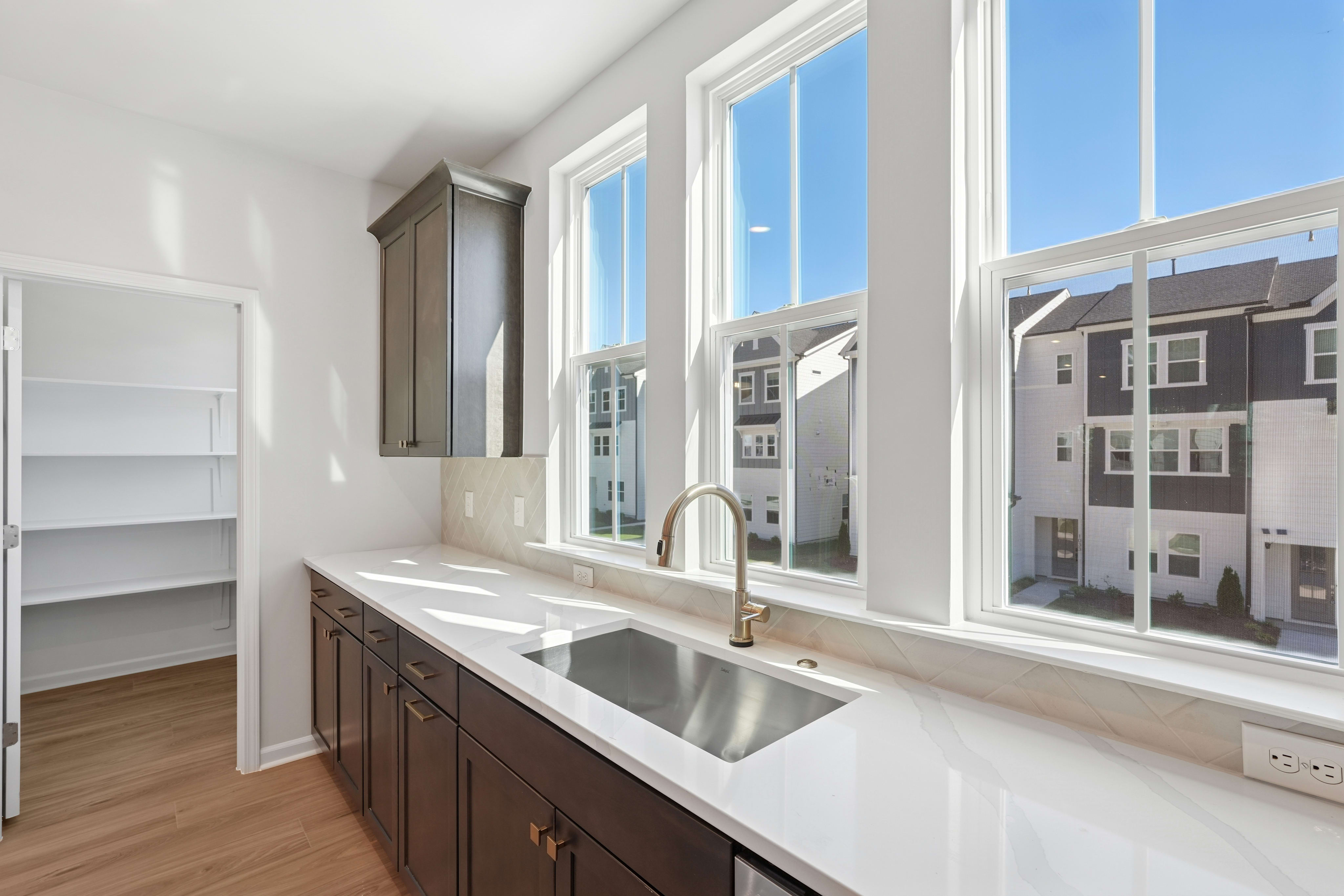 A modern kitchen with large windows overlooking a cityscape, featuring dark wood cabinets, a stainless steel sink, and a bright, airy atmosphere.
