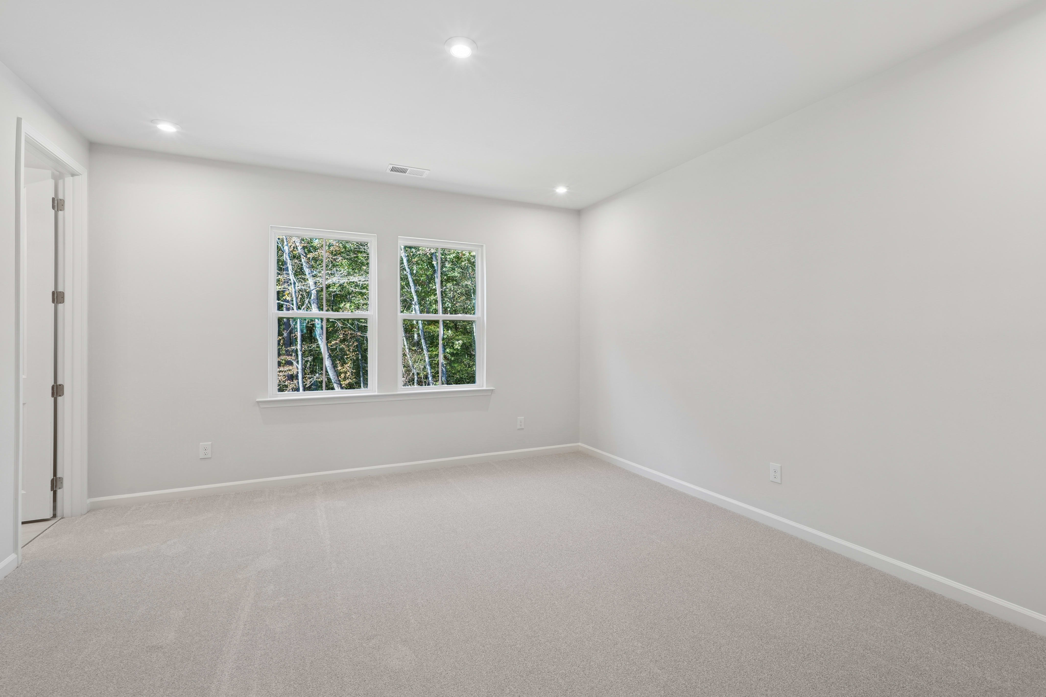 A bright, empty room with white walls, a hardwood floor, and large windows overlooking a wooded area outside.