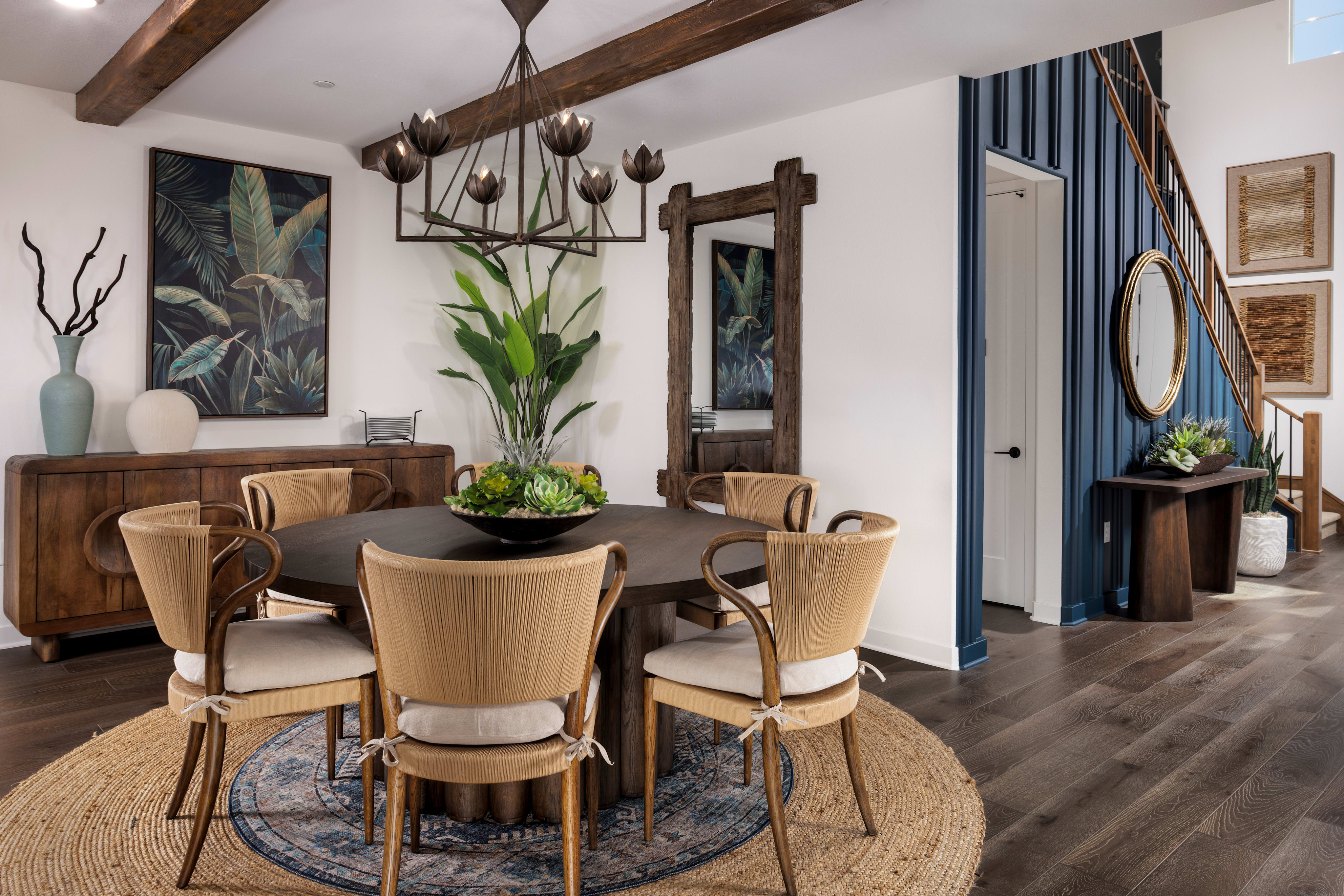 A cozy and stylish dining room with a wooden table surrounded by beige chairs, featuring a large tropical plant artwork, wooden beams, and a mirror on the wall.