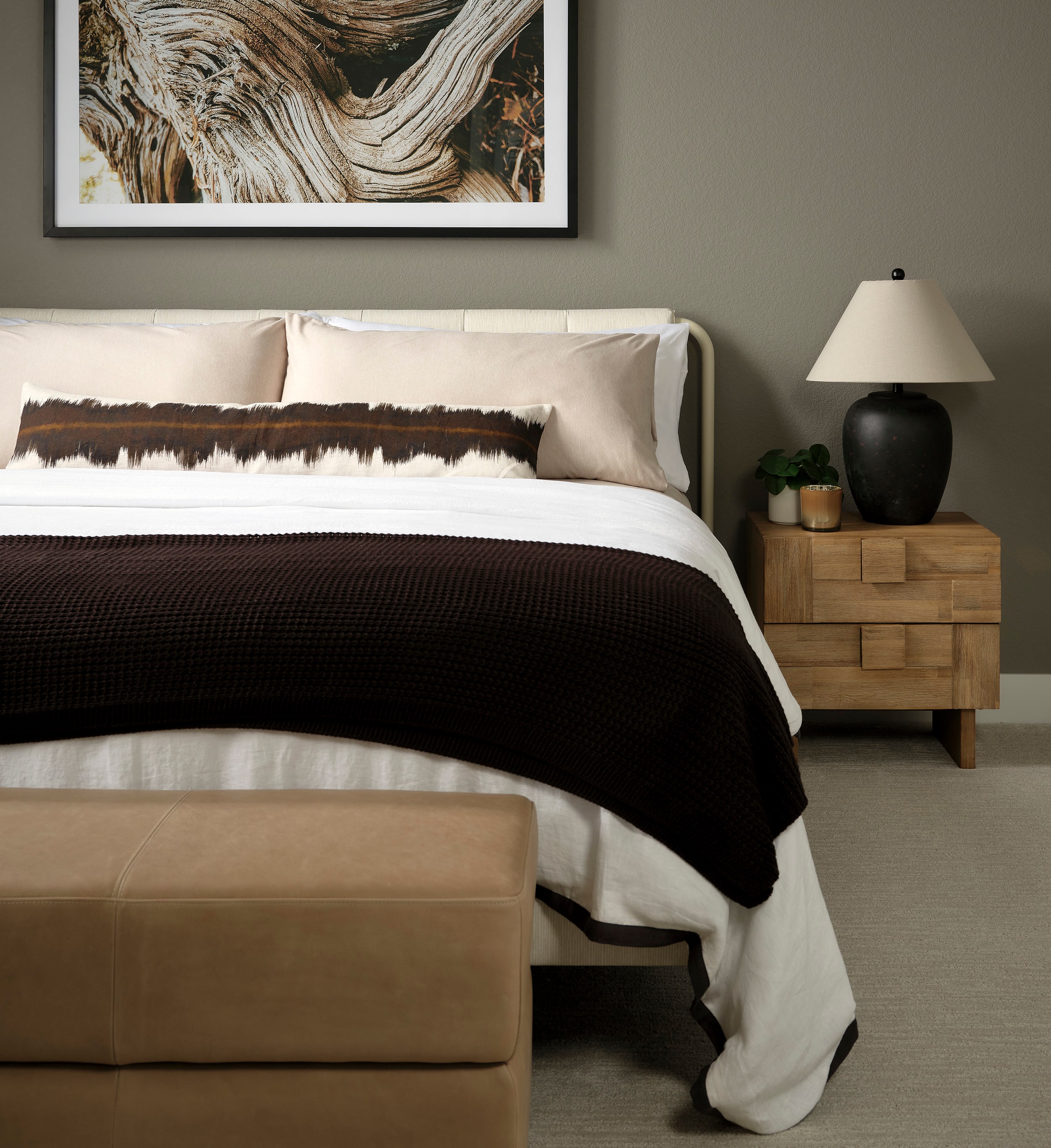 A cozy and inviting bedroom with a large framed artwork depicting abstract, swirling patterns on the wall, a plush bed with a dark blanket, and a wooden nightstand with a lamp and potted plant.