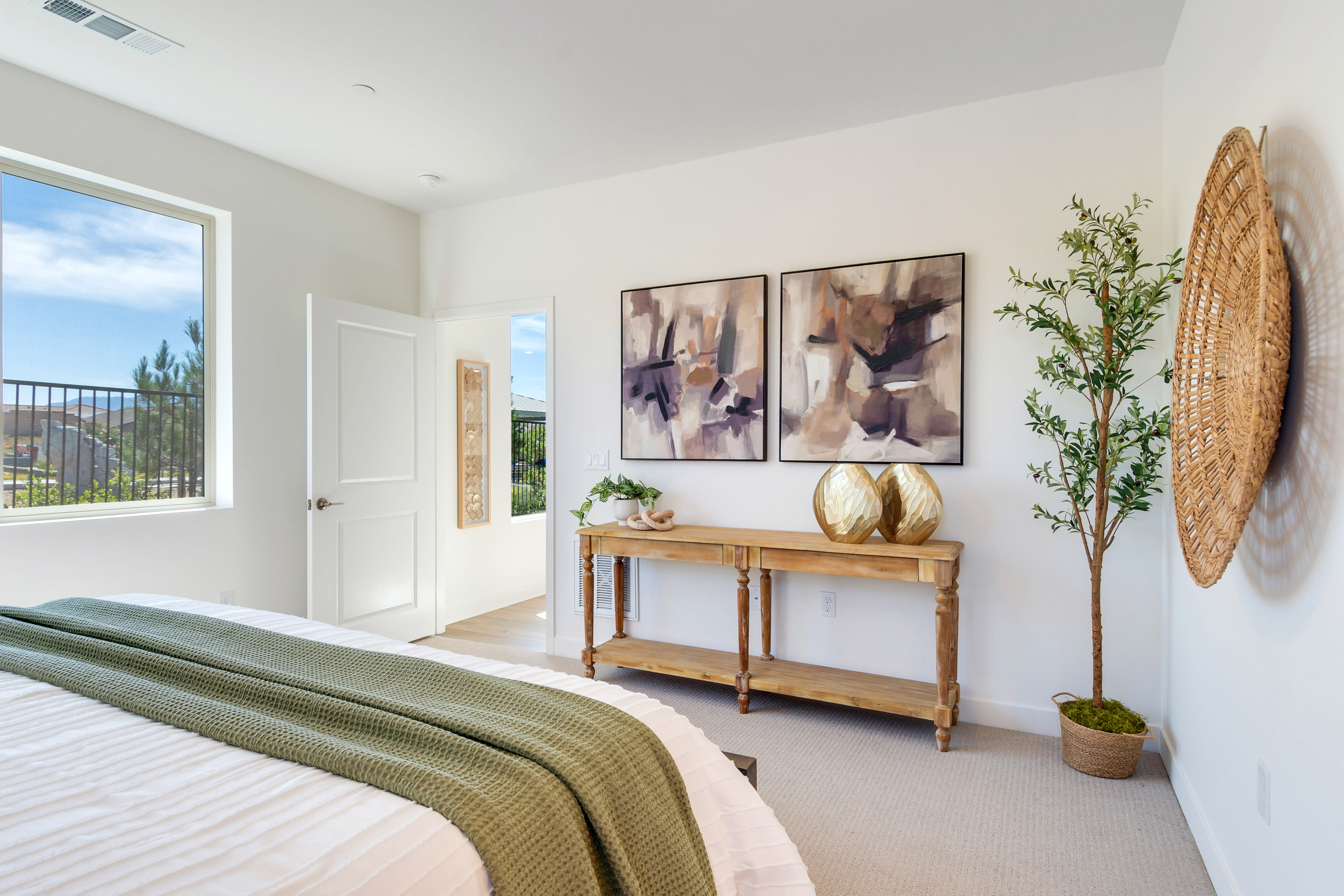 A cozy and inviting bedroom with a wooden console table, abstract artwork, and potted plants, set against a backdrop of a large window overlooking a scenic outdoor view.
