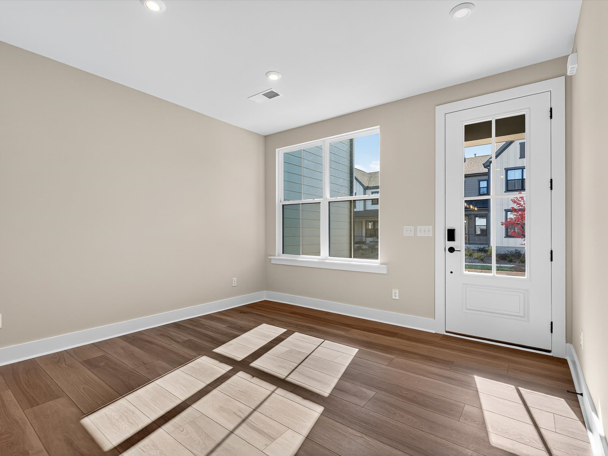 A spacious and well-lit room with a wooden floor, a large window, and a white door leading to the exterior.
