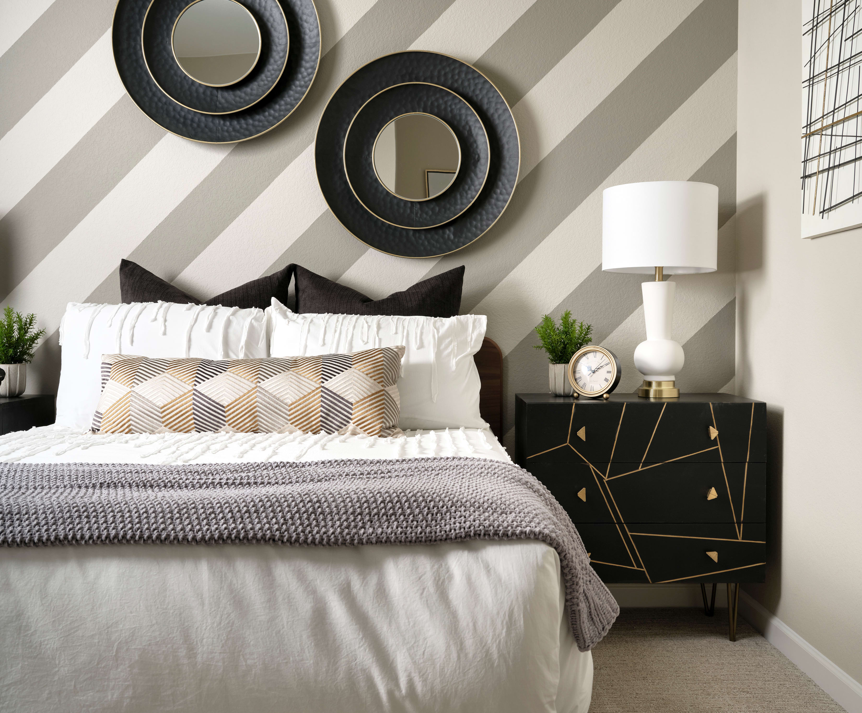 The image depicts a modern and stylish bedroom interior, featuring a large bed with a gray and white striped wall behind it, accented by two circular black and white wall decor pieces. A nightstand with a lamp and other decorative elements can be seen in the foreground.