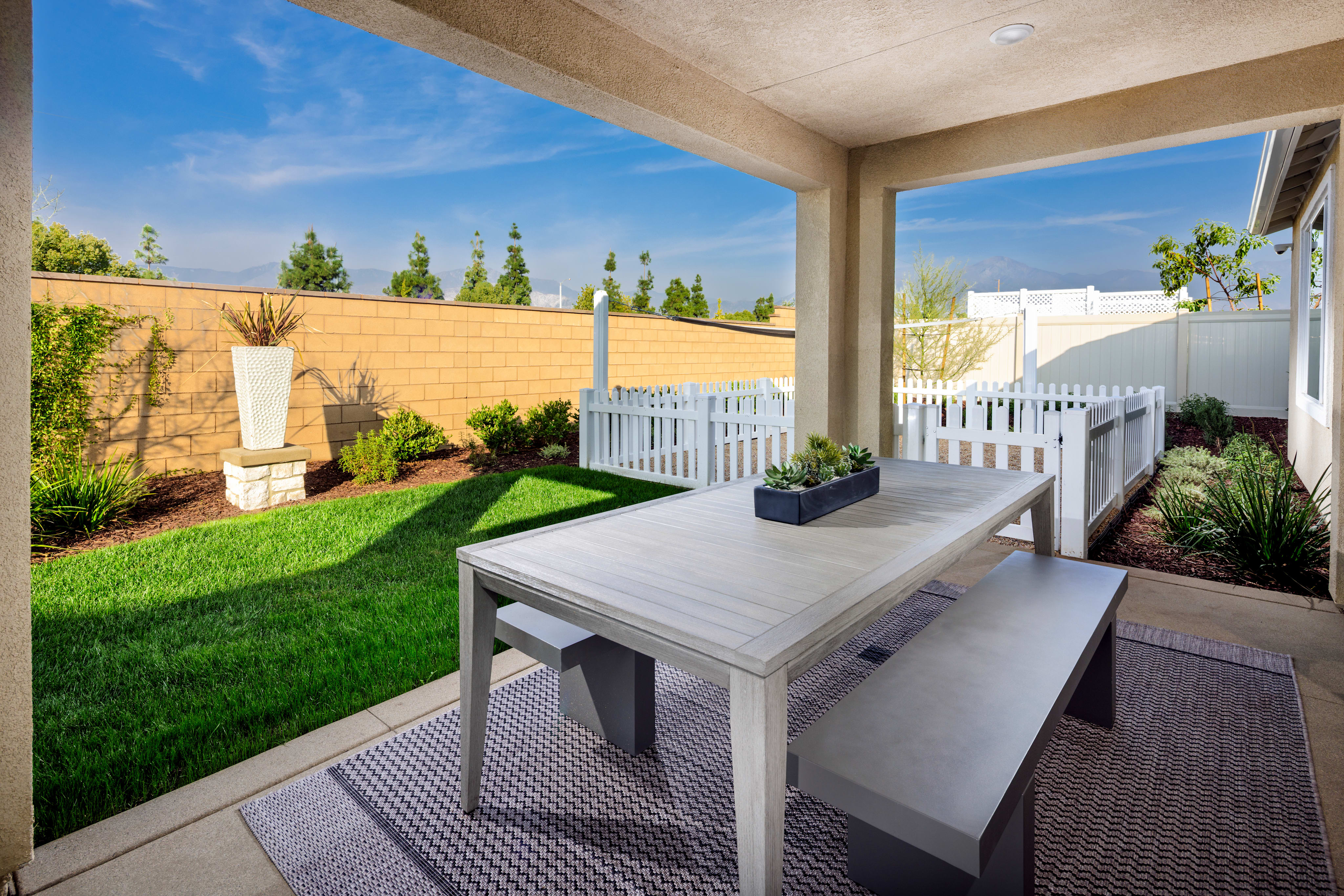 A well-designed outdoor patio area with a wooden table, lush green lawn, and a backdrop of a white fence and tall trees against a blue sky.