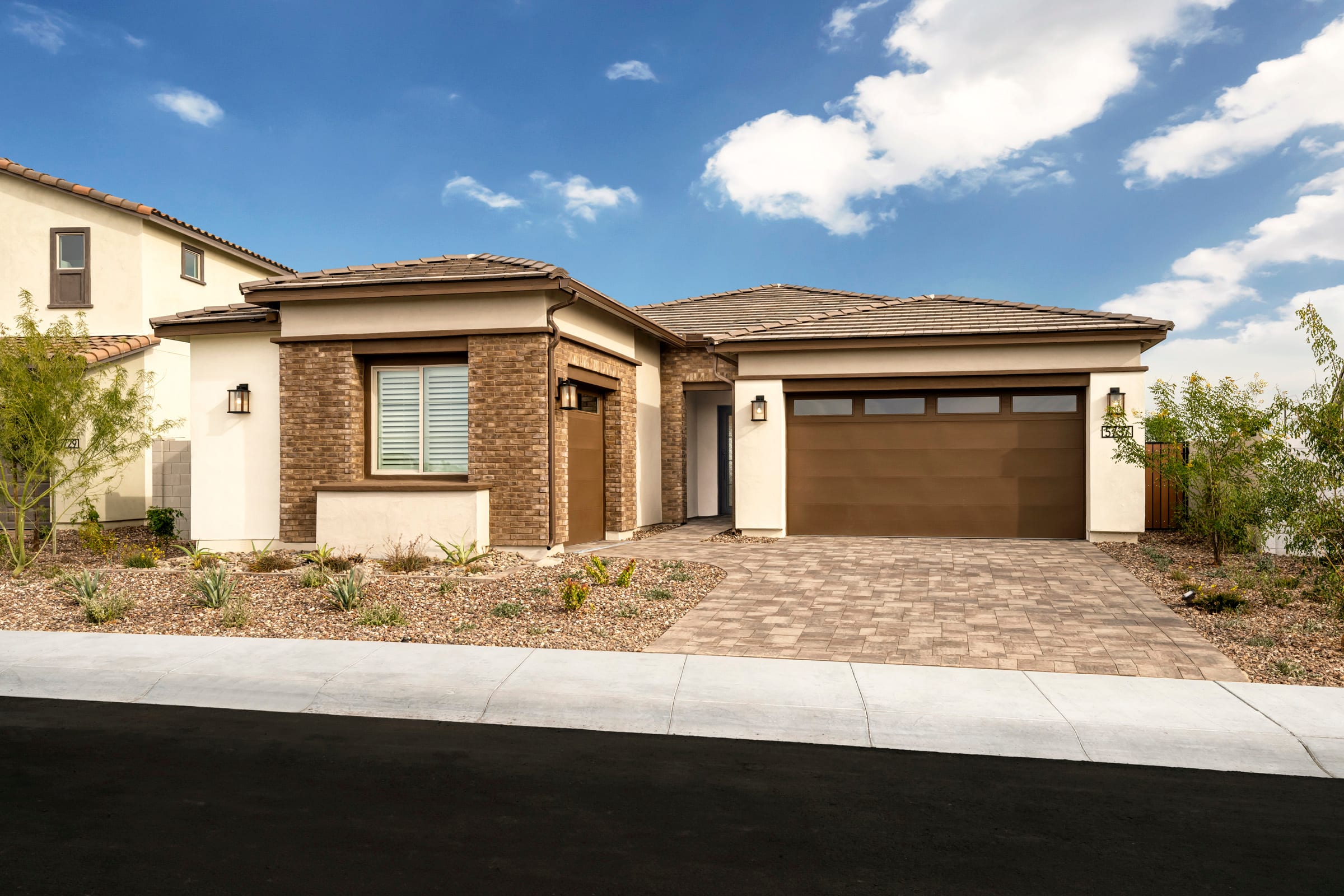 A single-story, modern-style house with a brick exterior, a tiled roof, and a two-car garage stands in a well-landscaped residential neighborhood against a backdrop of a blue sky with fluffy white clouds.